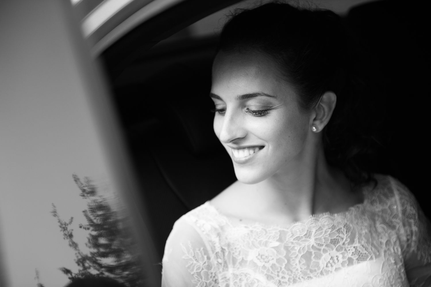 Black and white portrait of a smiling bride in a lace dress, looking down gracefully near a car window.
