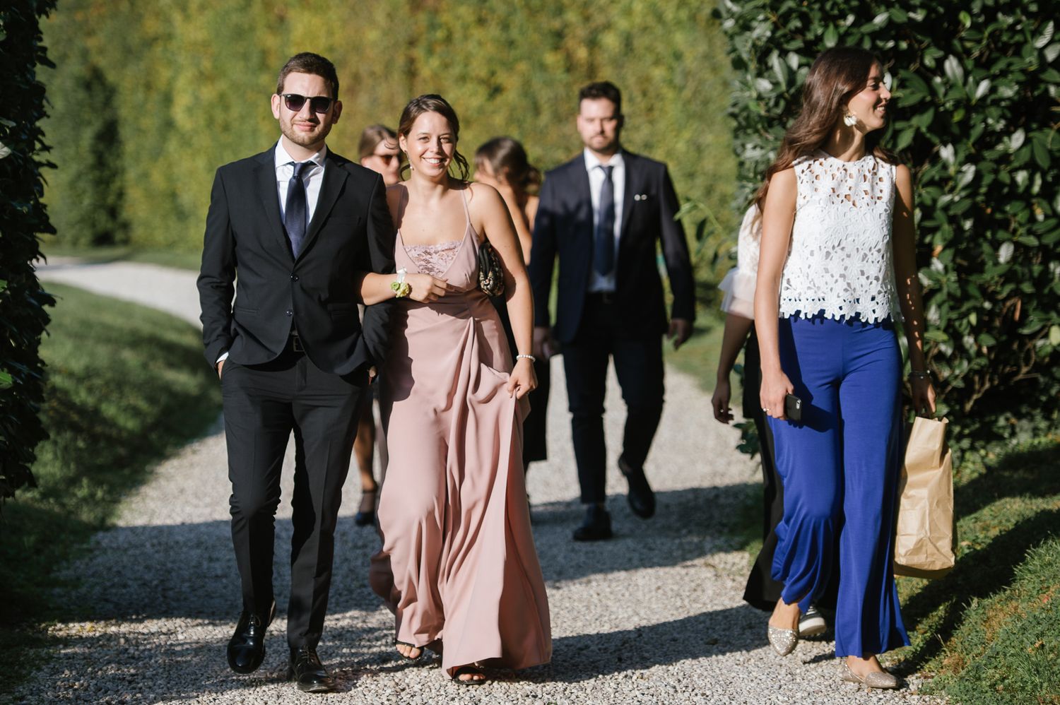 Wedding guests in elegant attire walking along garden path lined with tall hedges at outdoor venue.