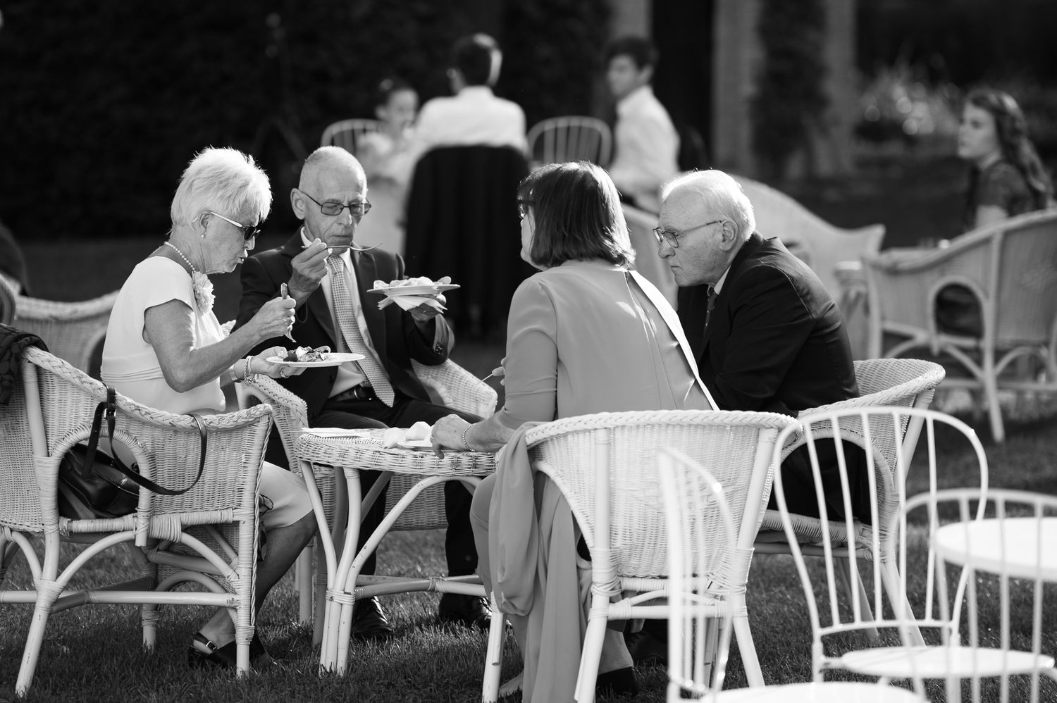Black and white photo of wedding guests enjoying food and conversation at outdoor garden reception tables.