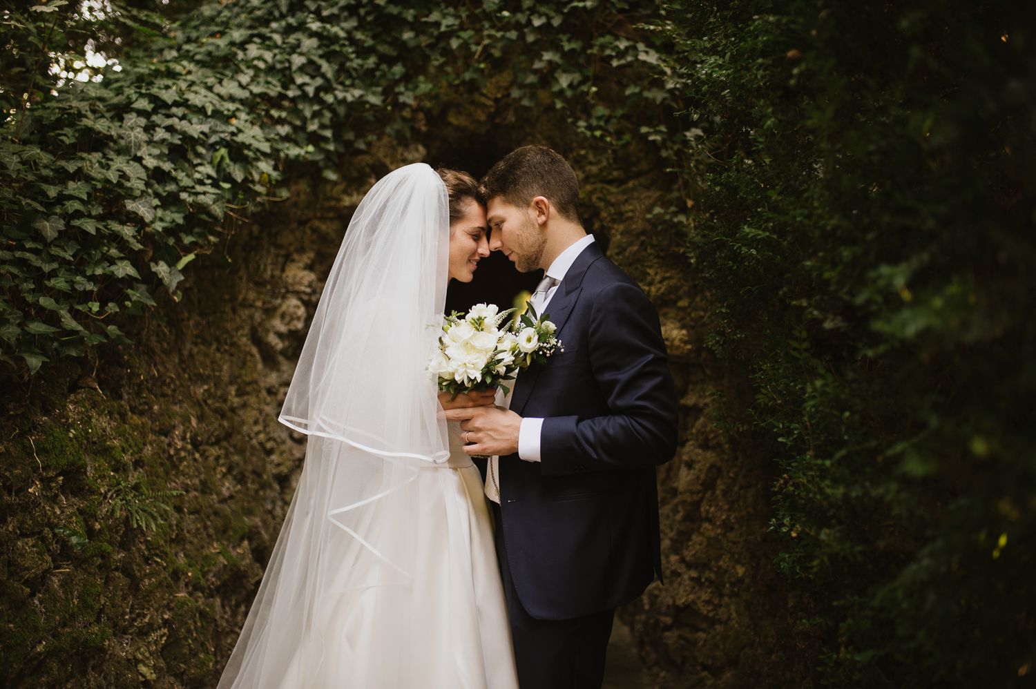 Newlywed couple sharing romantic moment touching foreheads in front of lush green ivy wall.