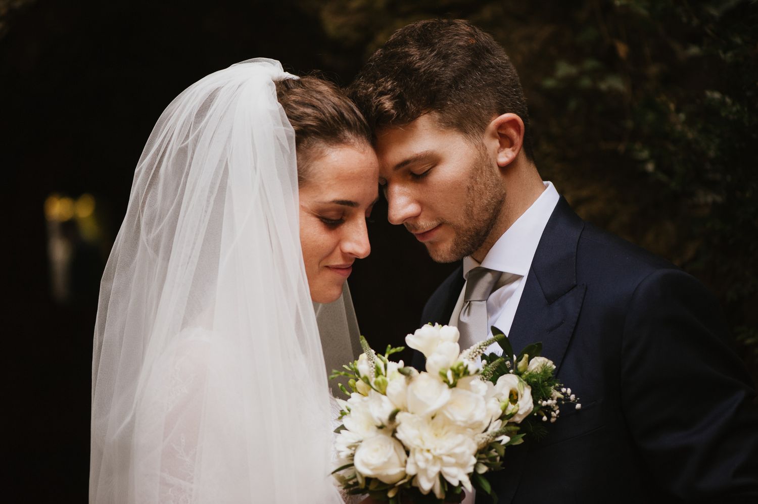 Romantic newlyweds touching foreheads tenderly with white bridal bouquet outdoors.