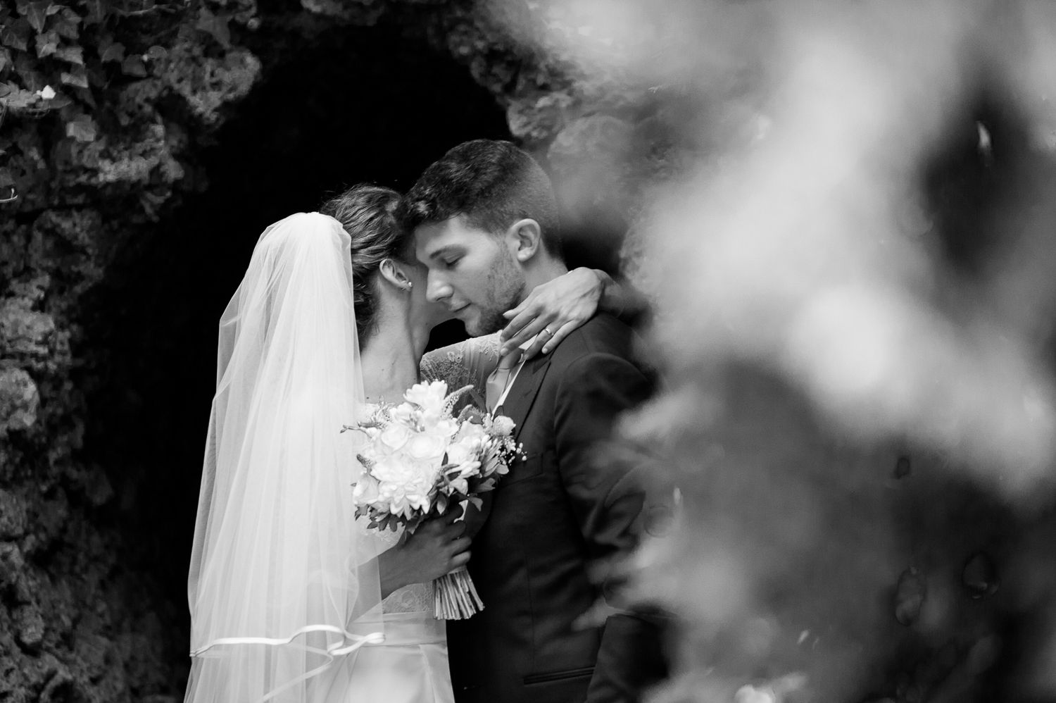 Black and white wedding photo of bride and groom sharing a romantic kiss under a stone arch.
