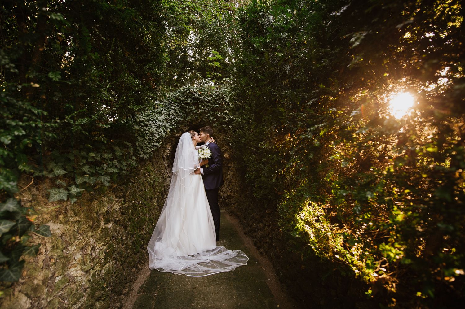 Bride and groom kissing in a sunlit ivy-covered garden pathway on wedding day.