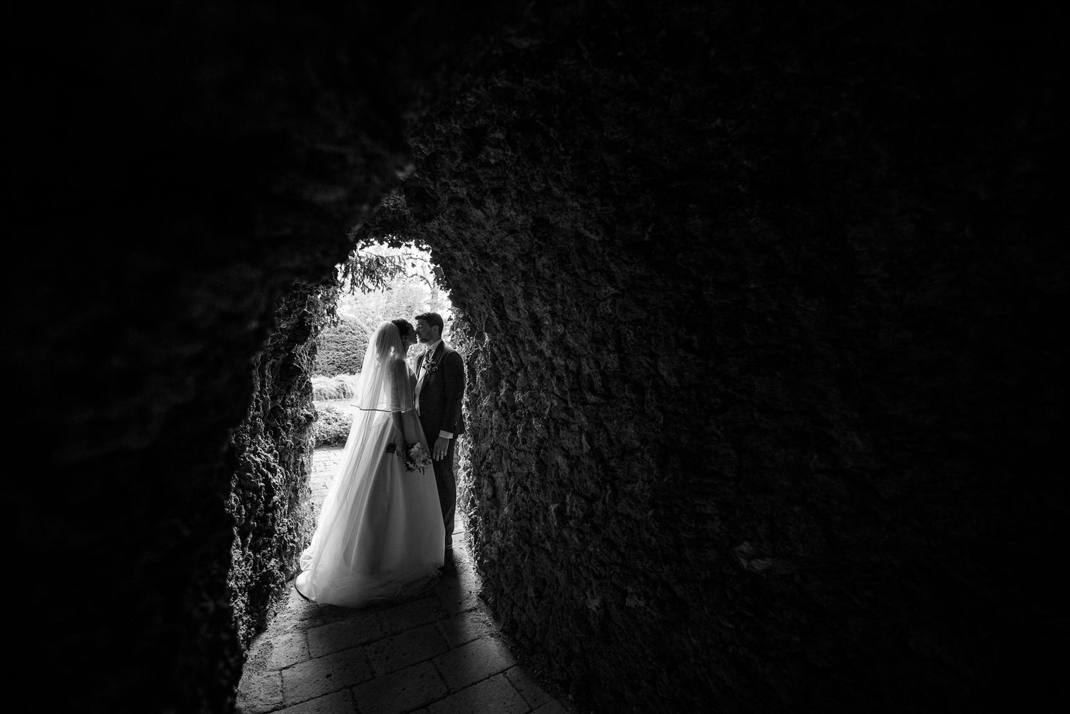 Black and white photo of bride and groom embracing in a stone tunnel archway on their wedding day.