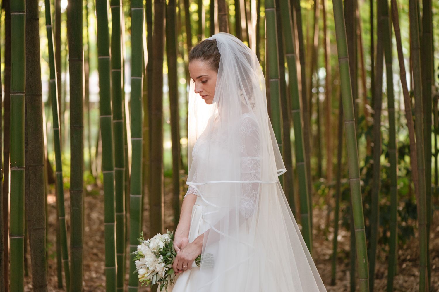 Elegant bride in white gown and veil holding bouquet among tall bamboo trees.