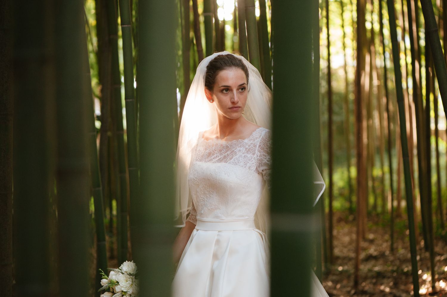 Elegant bride in lace wedding gown and veil stands among tall bamboo trees holding a bouquet.