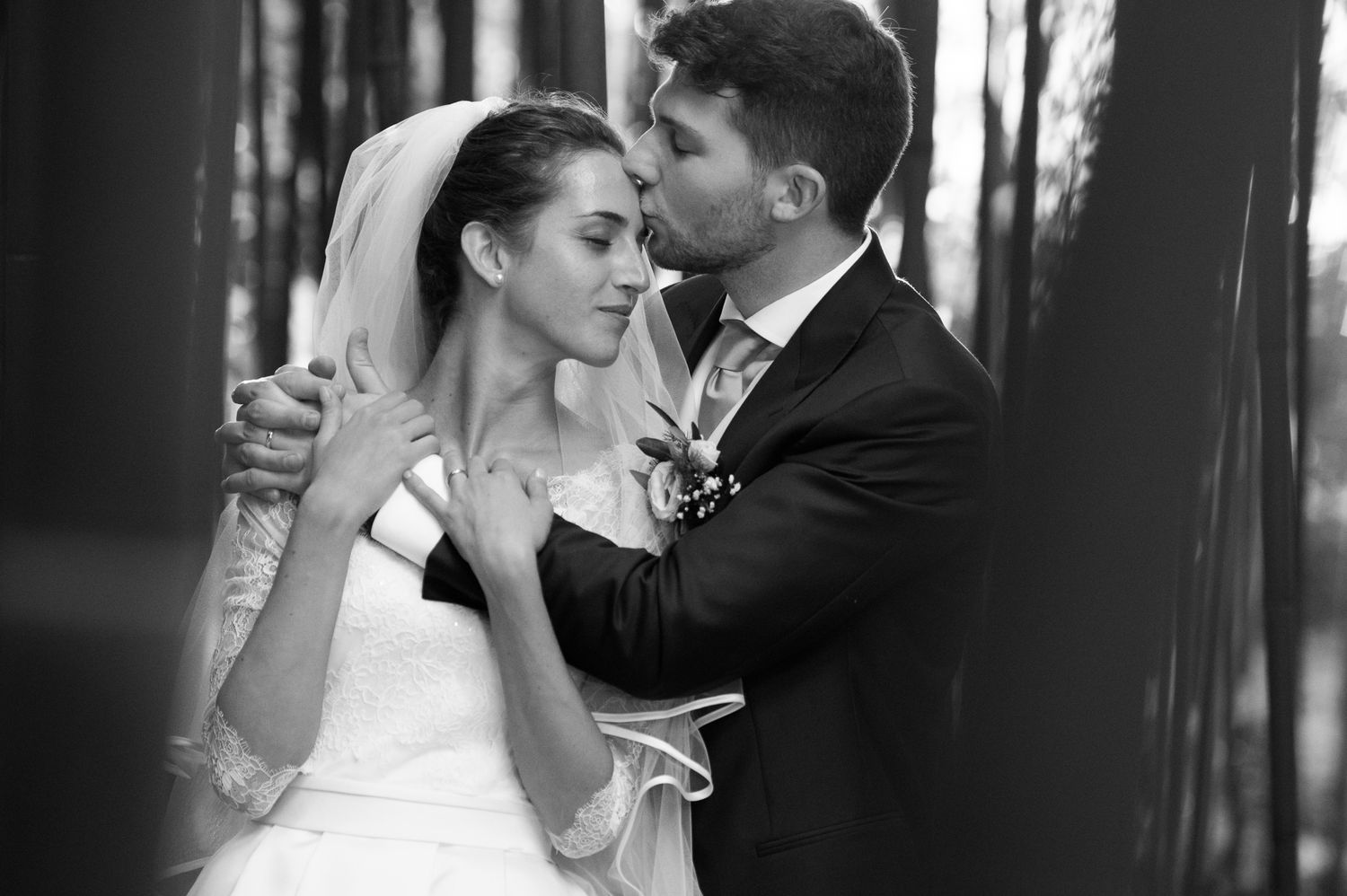 Black and white wedding photo of bride and groom embracing tenderly among trees.