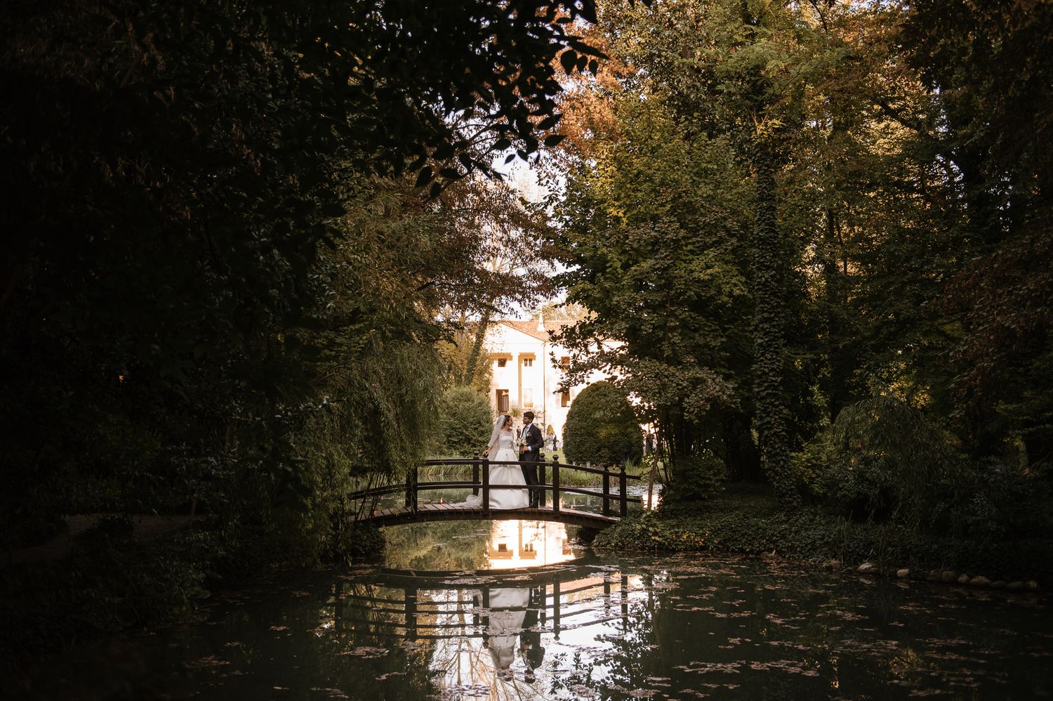 Newlyweds share a romantic moment on a wooden bridge reflected in a tranquil garden pond.