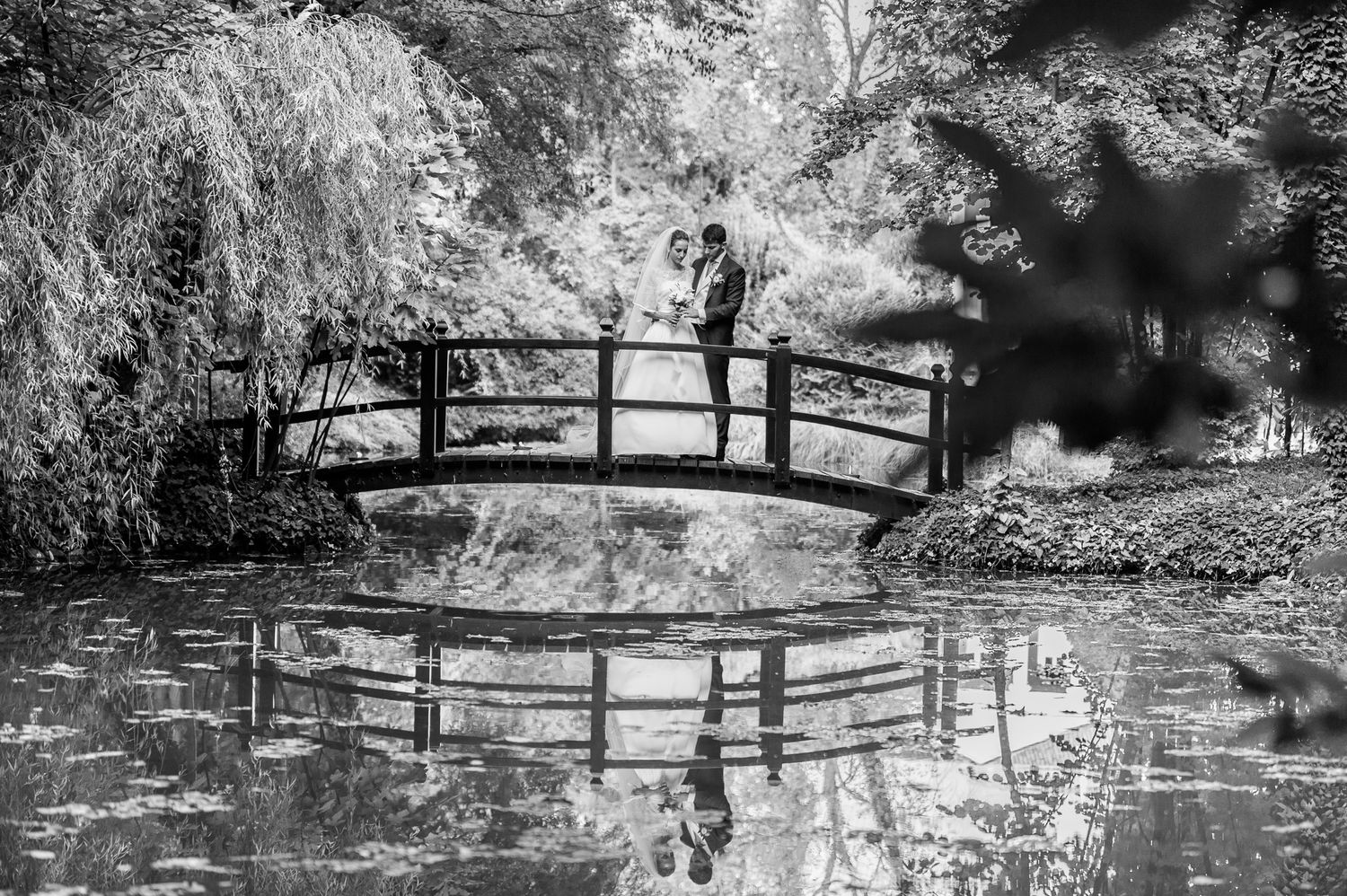A bride and groom stand on an arched bridge reflected in a tranquil pond, captured in black and white.