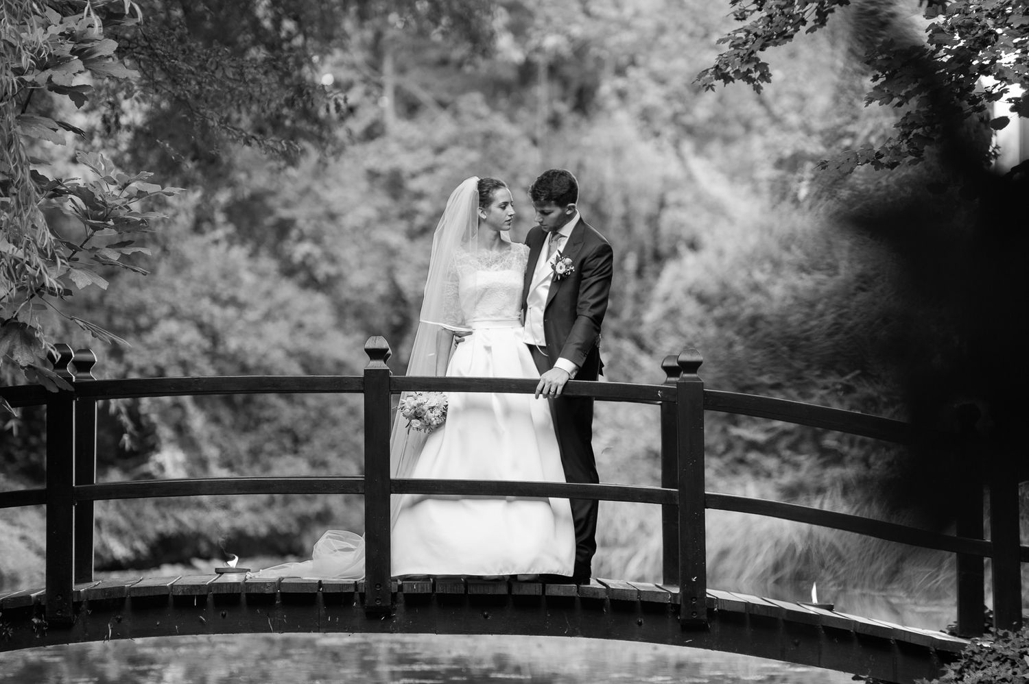 Bride and groom standing on a wooden bridge surrounded by lush greenery in black and white.