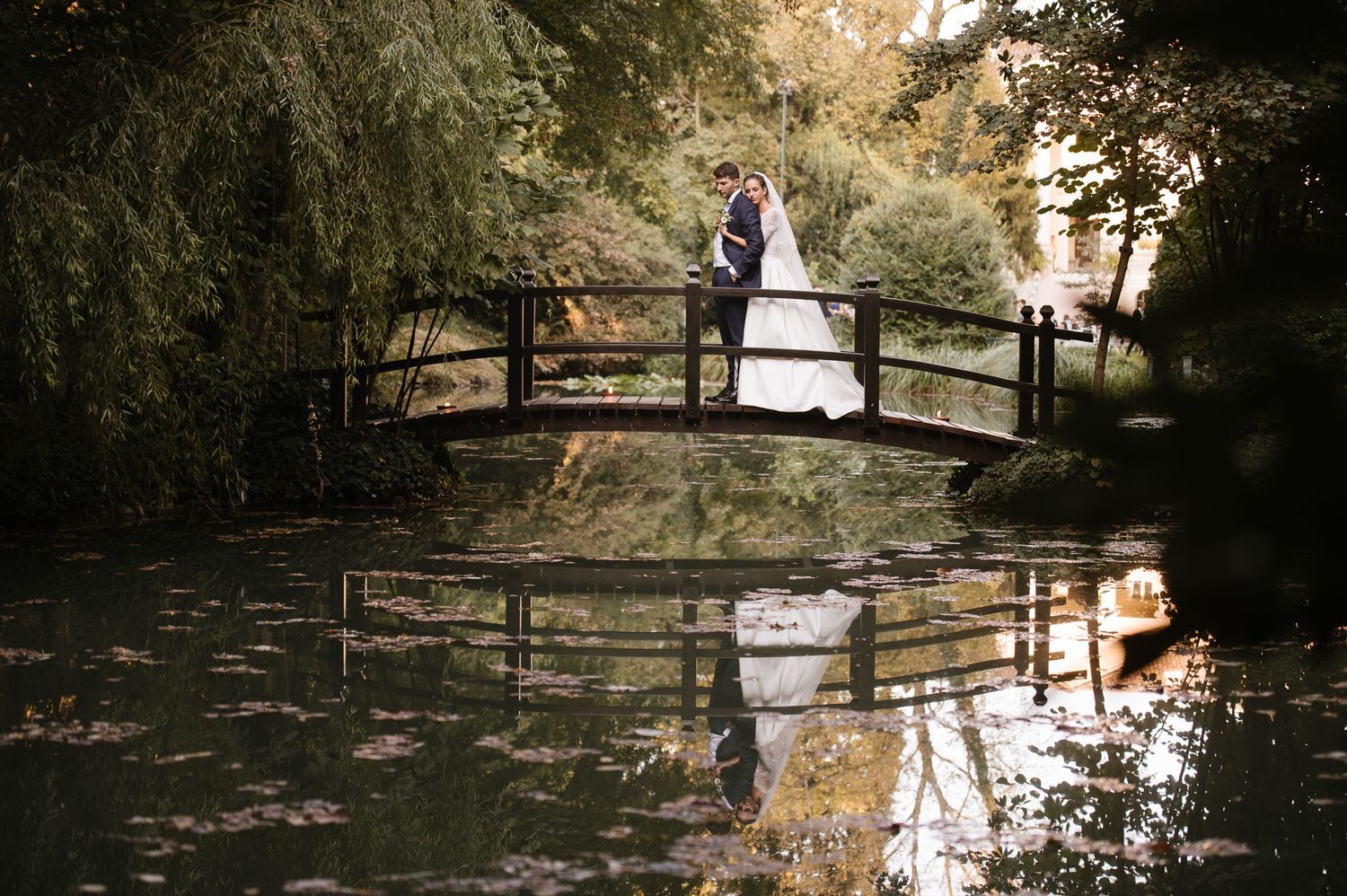 Wedding couple standing on a wooden bridge reflected in a calm pond surrounded by trees.