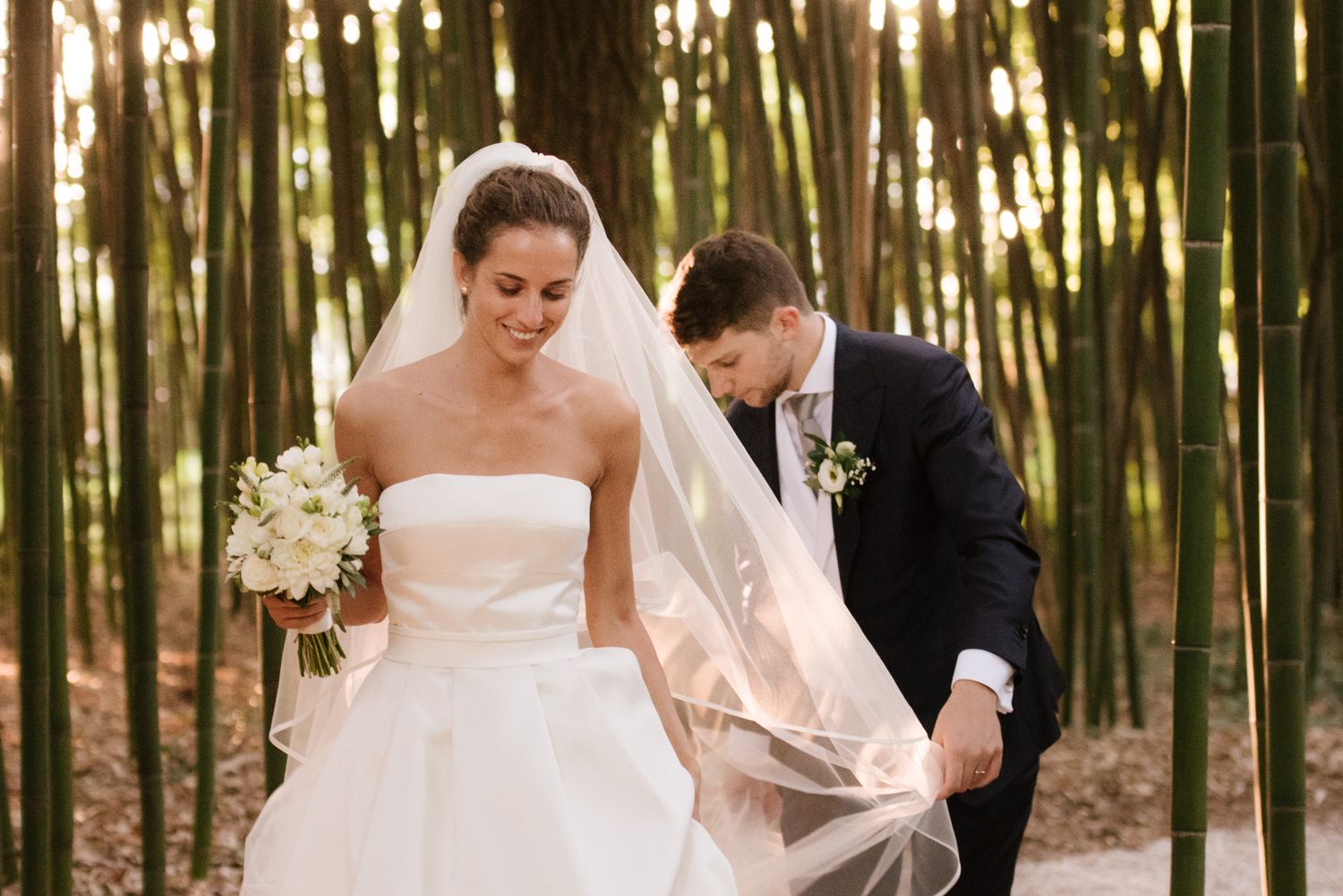 Bride in strapless gown holding bouquet while groom adjusts veil in bamboo forest.