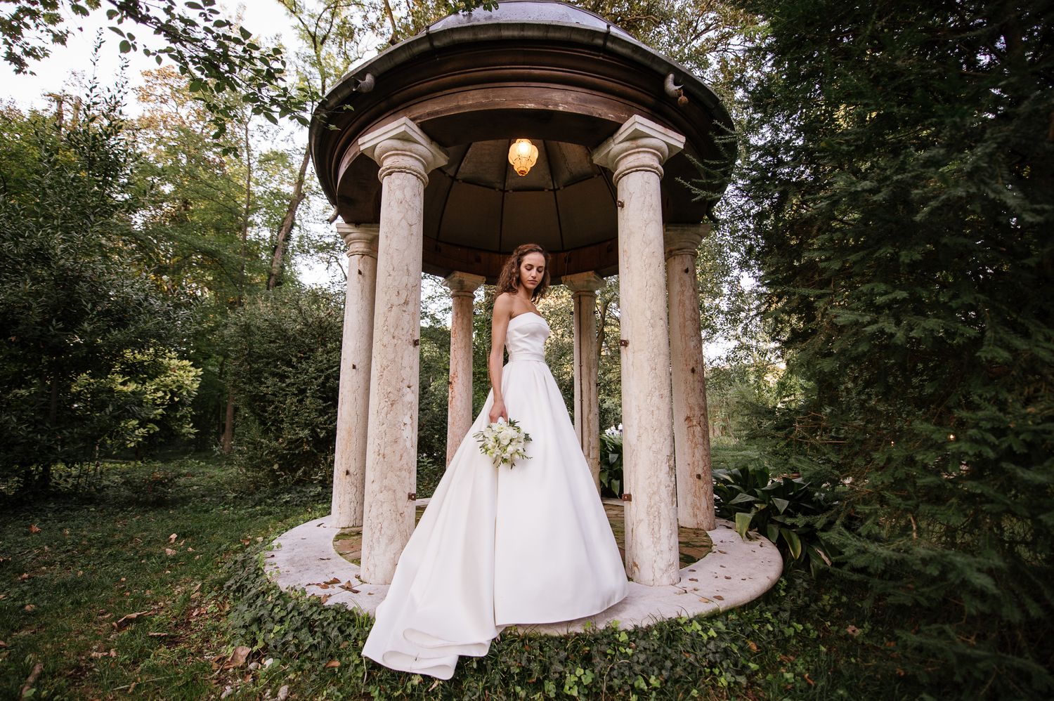 Elegant bride in white ball gown standing under a stone gazebo surrounded by lush green trees.