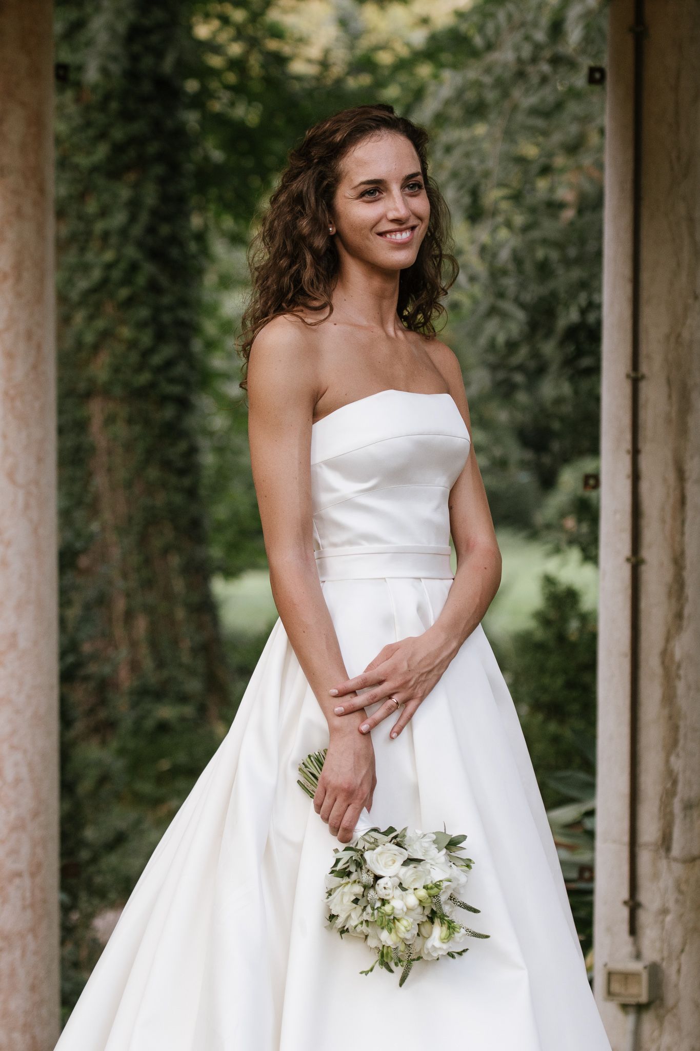 Smiling bride in strapless white satin gown holding white floral bouquet outdoors near stone columns.