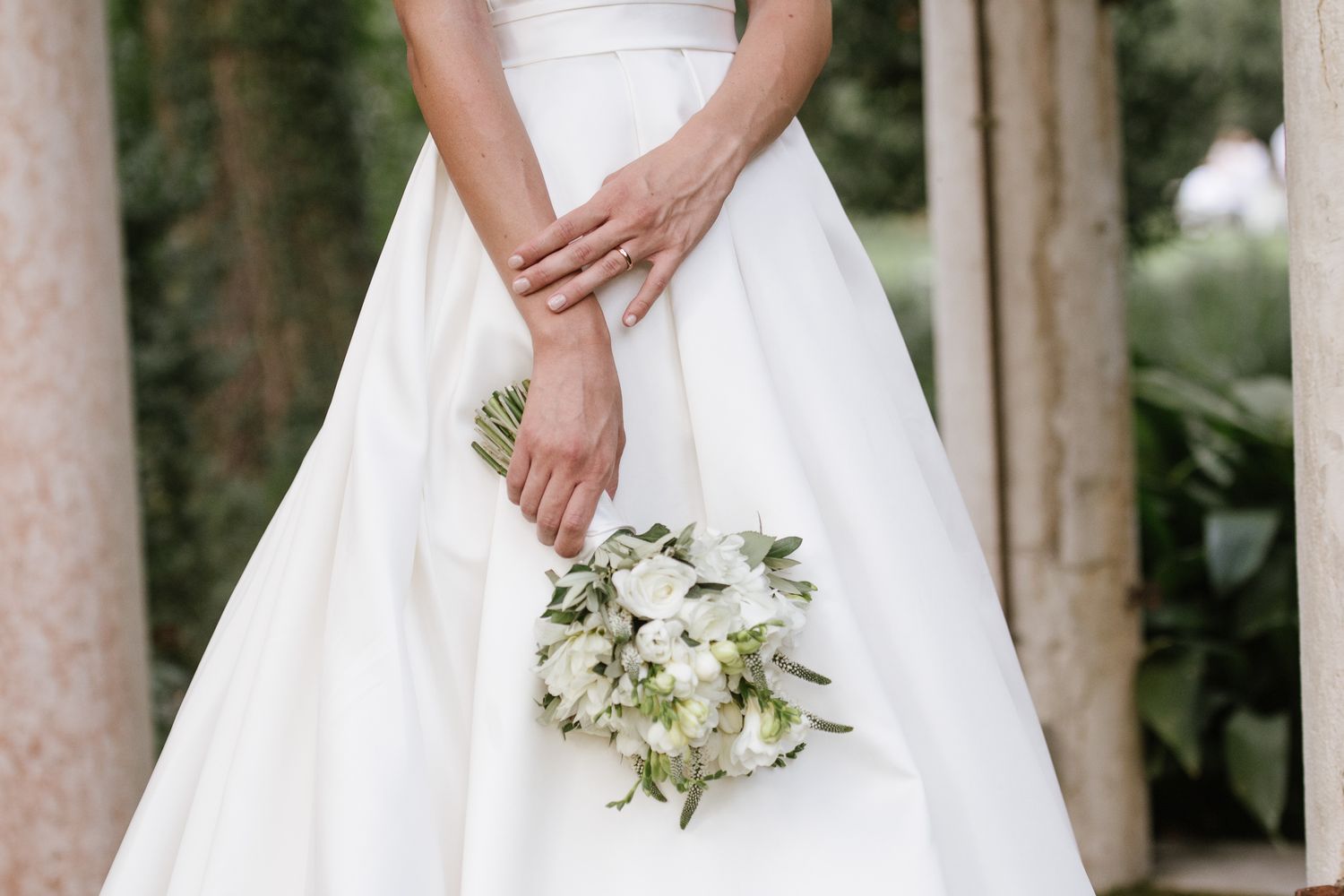 Close-up of bride in white wedding dress holding a white floral bouquet with greenery.