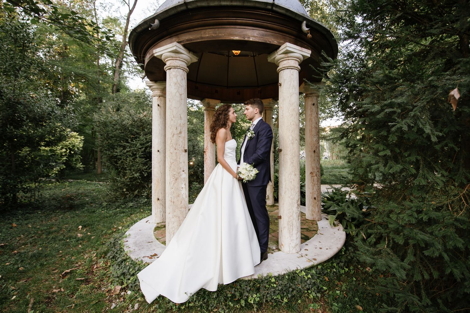 Bride and groom share a romantic moment under a stone gazebo surrounded by lush greenery.