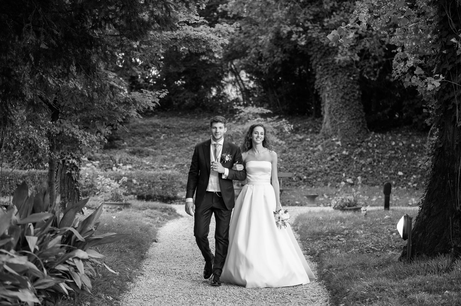 Black and white photo of a bride and groom walking hand in hand along a garden path lined with trees.