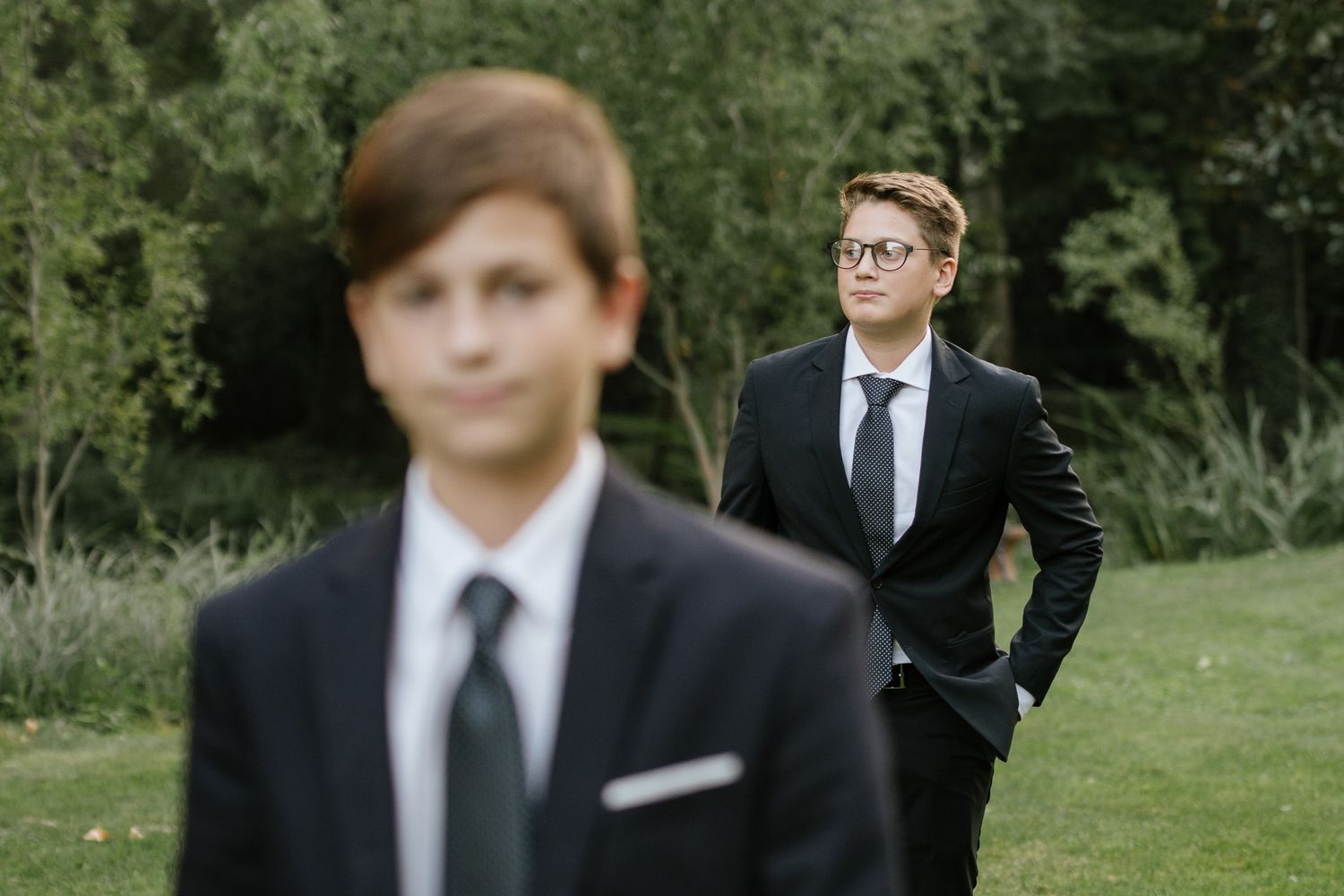 Two young men in dark suits with ties standing on a green lawn at an outdoor event.
