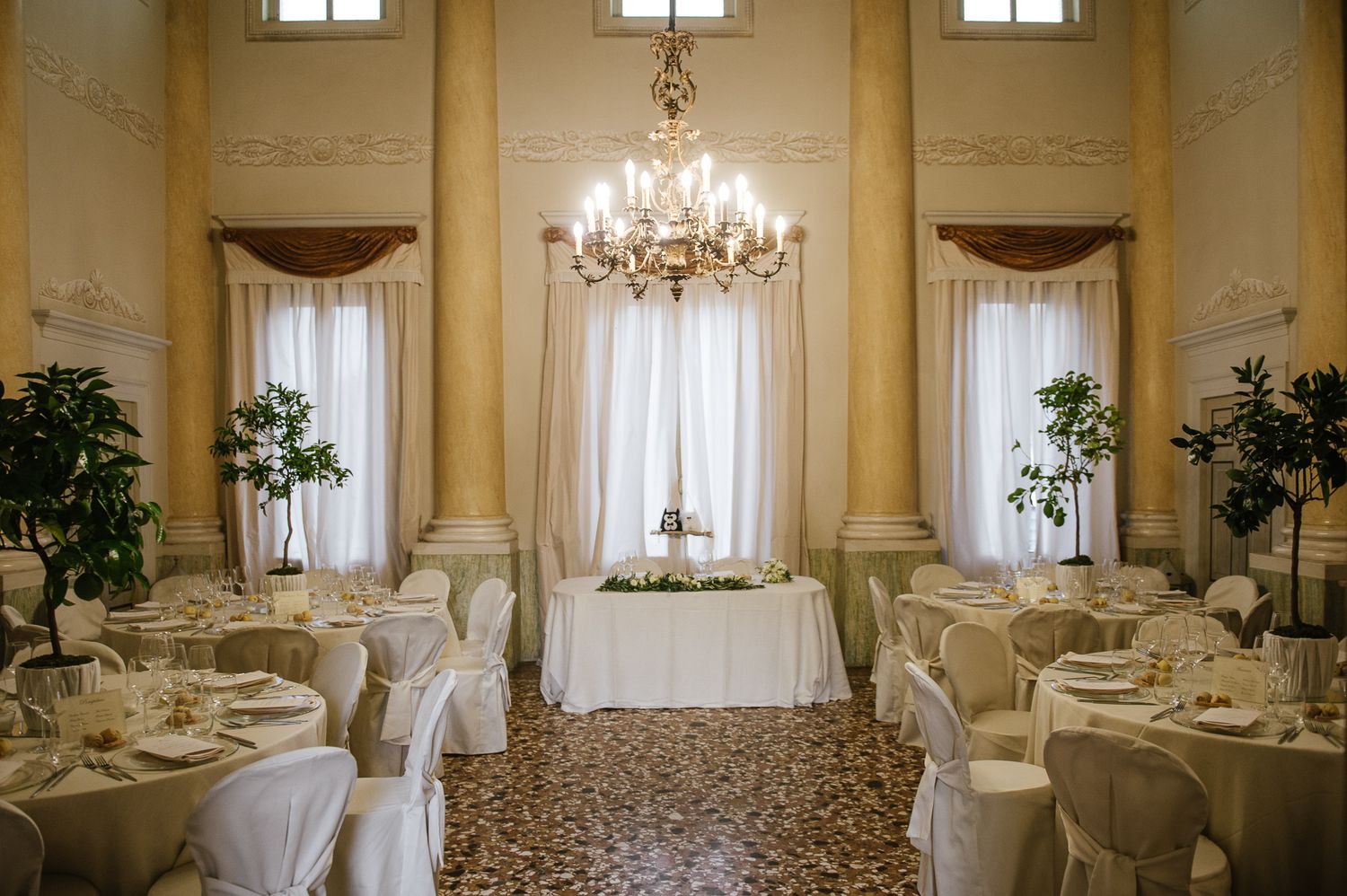 An elegant wedding reception hall features chandelier lighting, white draped tables, and tall floral centerpieces.