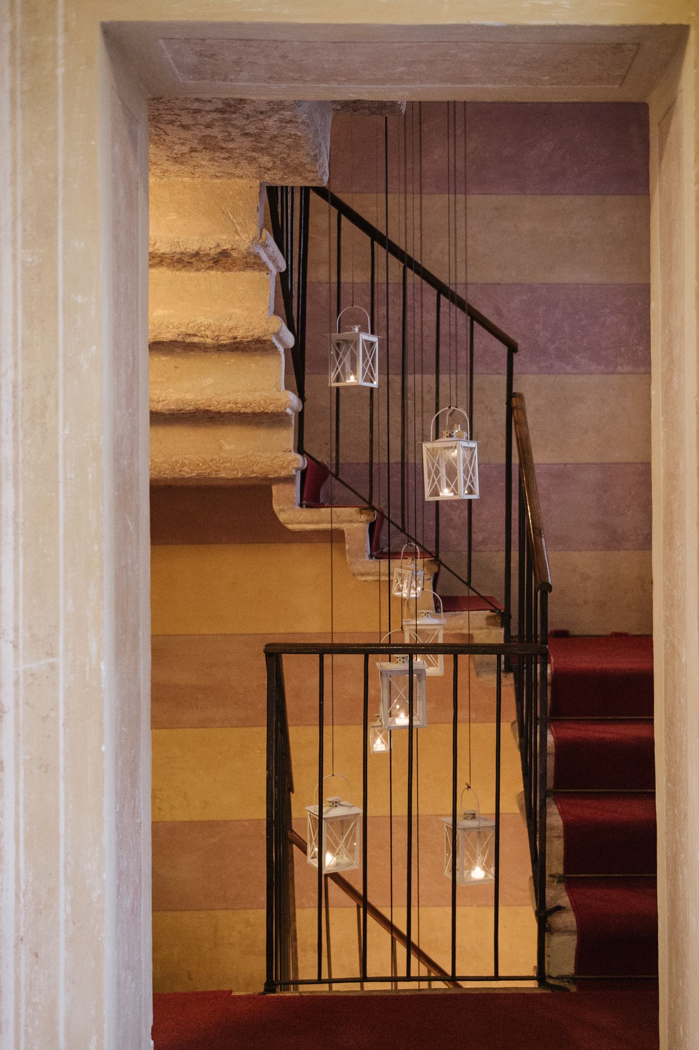 Elegant staircase with hanging lanterns and red carpet steps, framed by colorful striped walls in a historic venue.