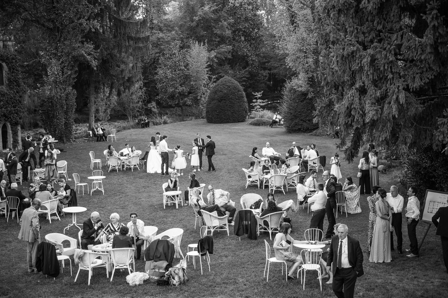 Black and white aerial view of an elegant outdoor wedding reception with guests mingling around white tables on a lush garden lawn.