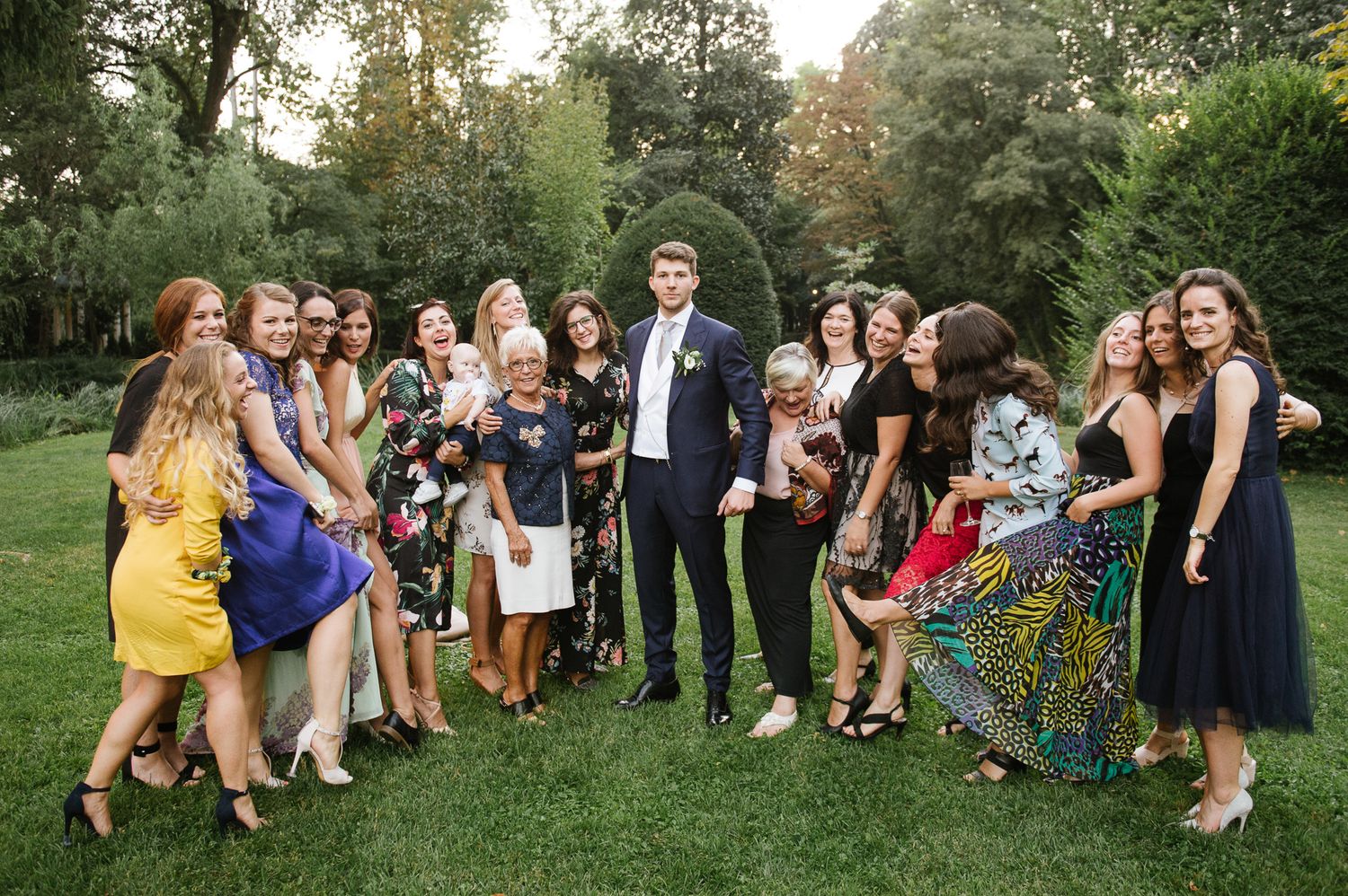 Groom in navy suit posing with large group of female wedding guests on lush green garden lawn.