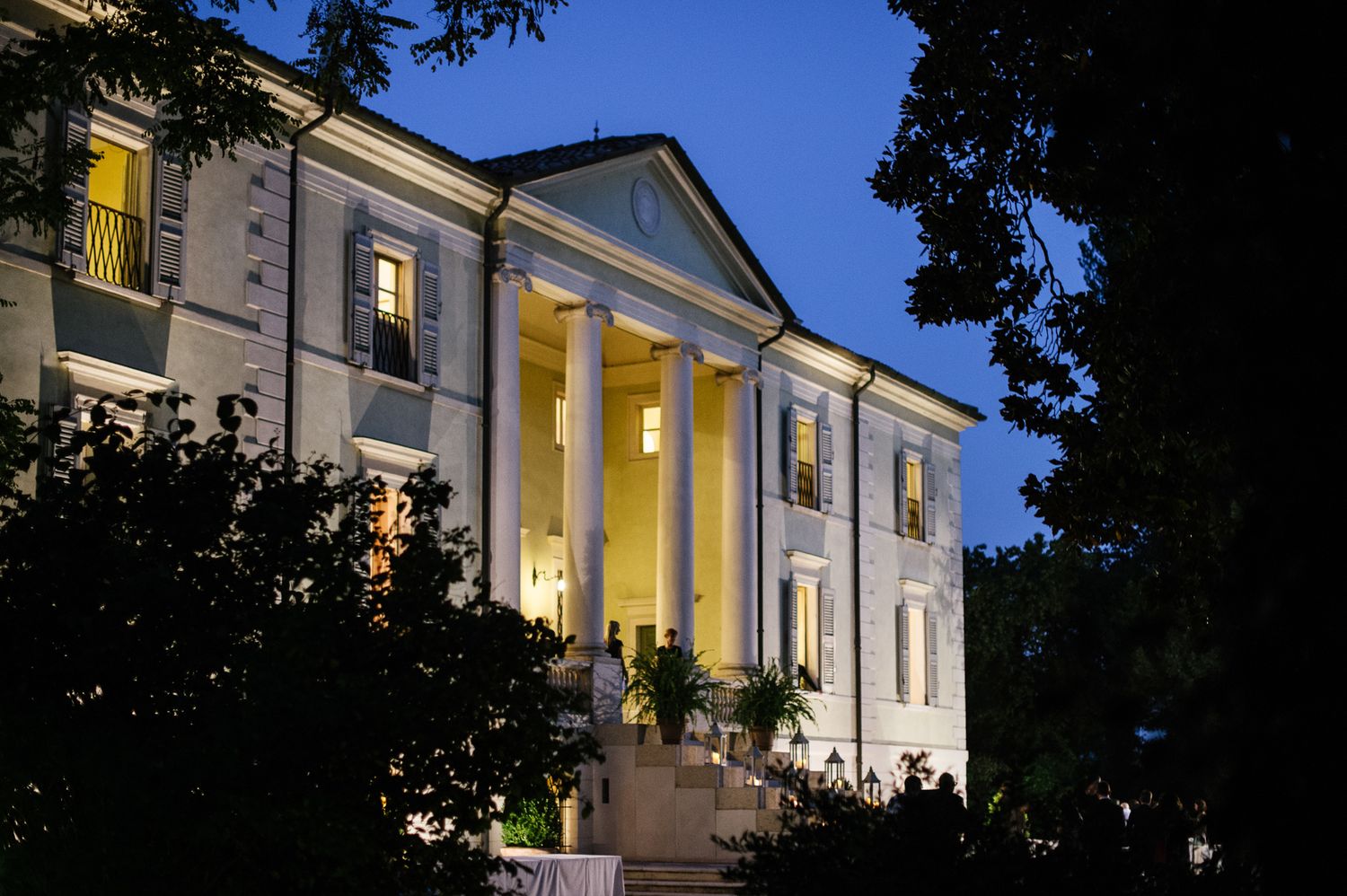 Illuminated neoclassical wedding venue with white columns glowing at dusk surrounded by lush trees.