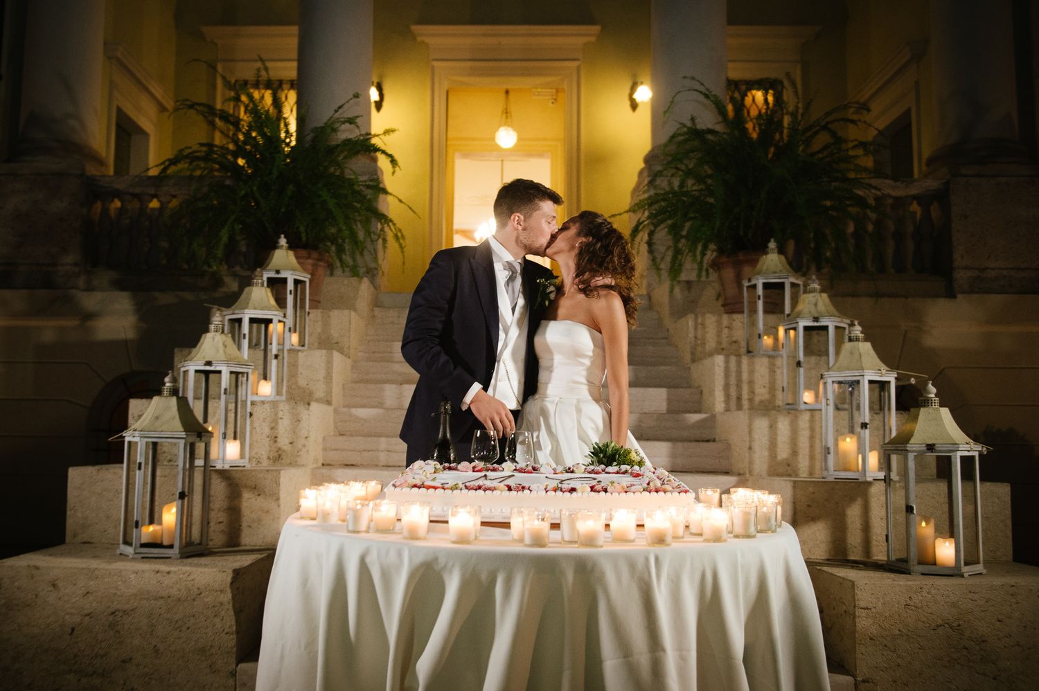 Bride and groom cutting wedding cake on candlelit steps of a grand villa at night.