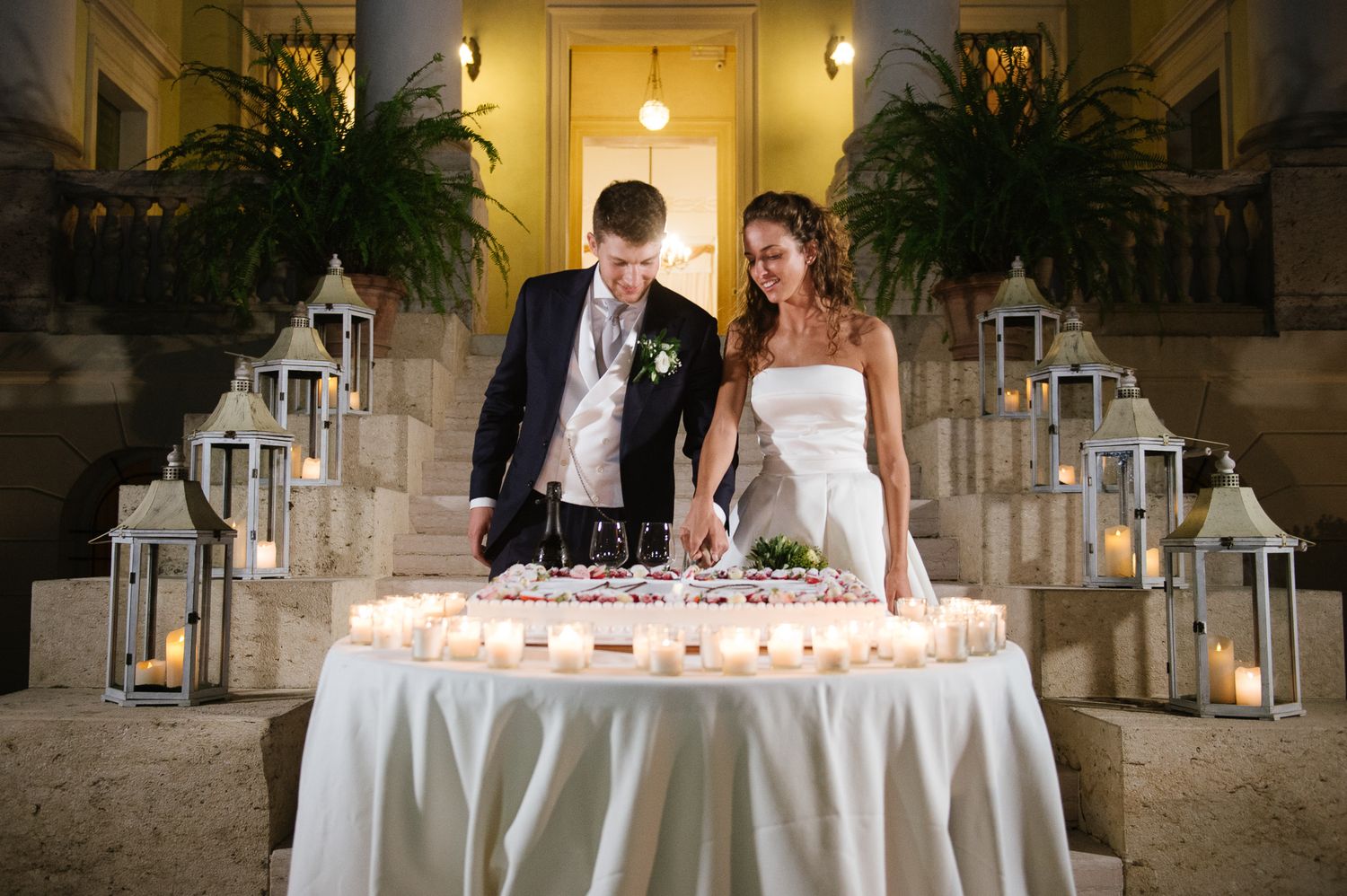 Bride and groom cutting wedding cake surrounded by glowing lanterns and candles at elegant evening reception.