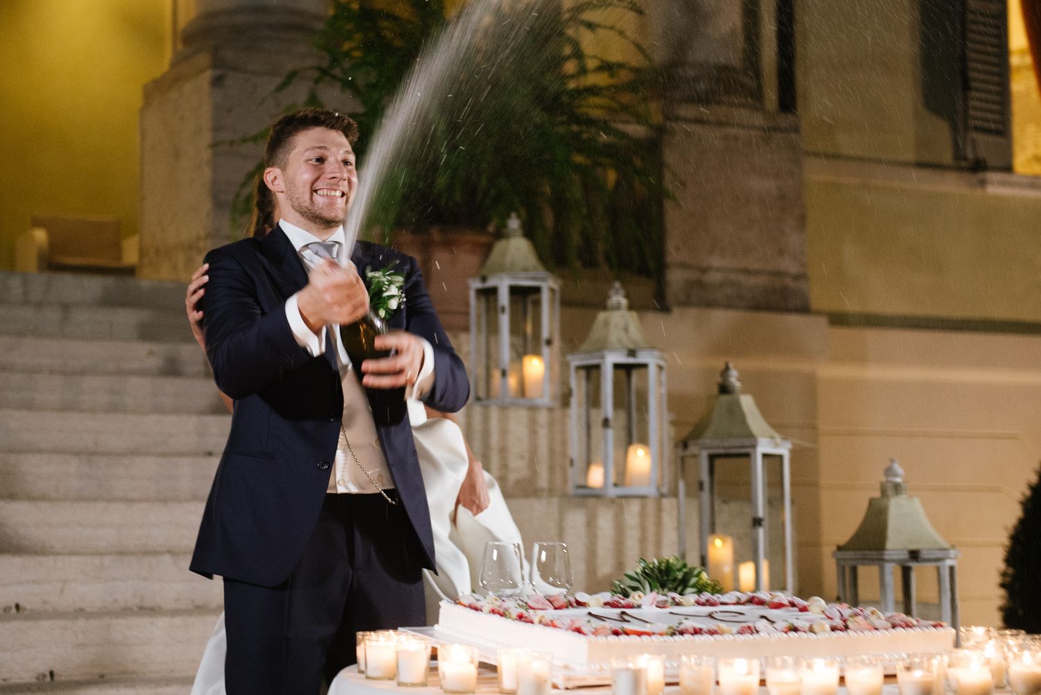 A groom pops a champagne bottle surrounded by candles and lanterns during an outdoor wedding celebration.