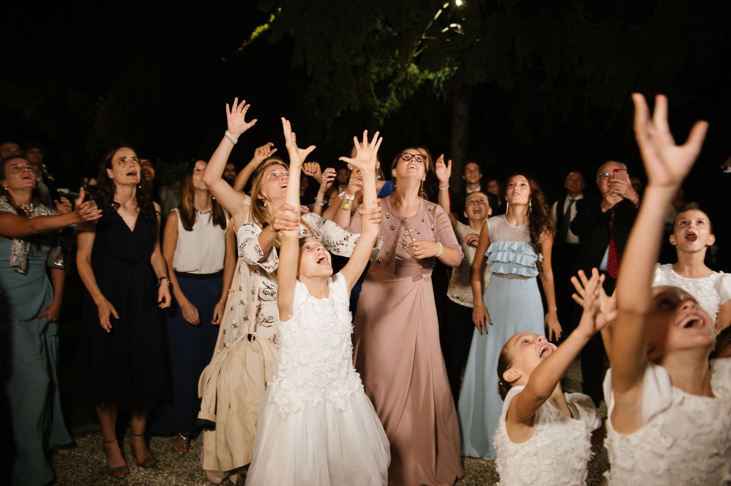 Wedding guests reaching up to catch the bridal bouquet toss at an outdoor evening reception.