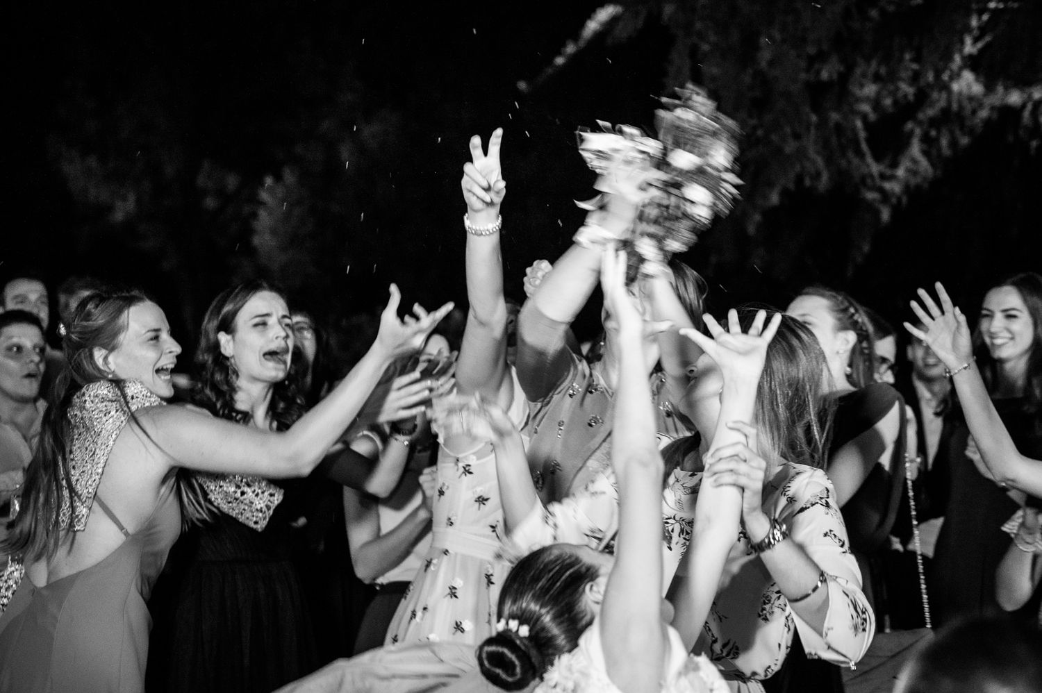 Black and white photo of wedding guests reaching up to catch the bridal bouquet toss at a reception.