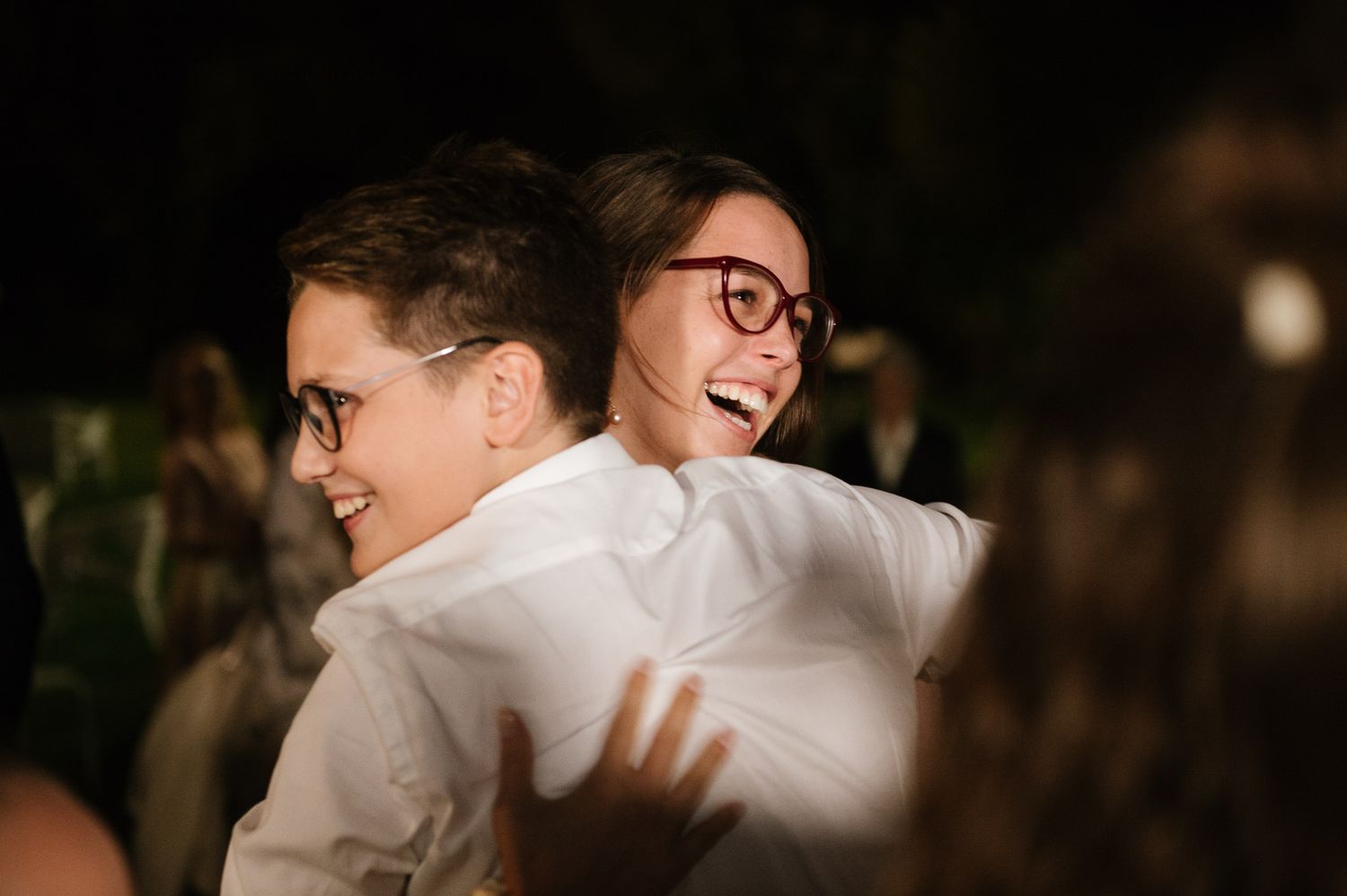 Two people sharing a joyful hug and laughing at an outdoor evening celebration, both wearing glasses and white shirts.