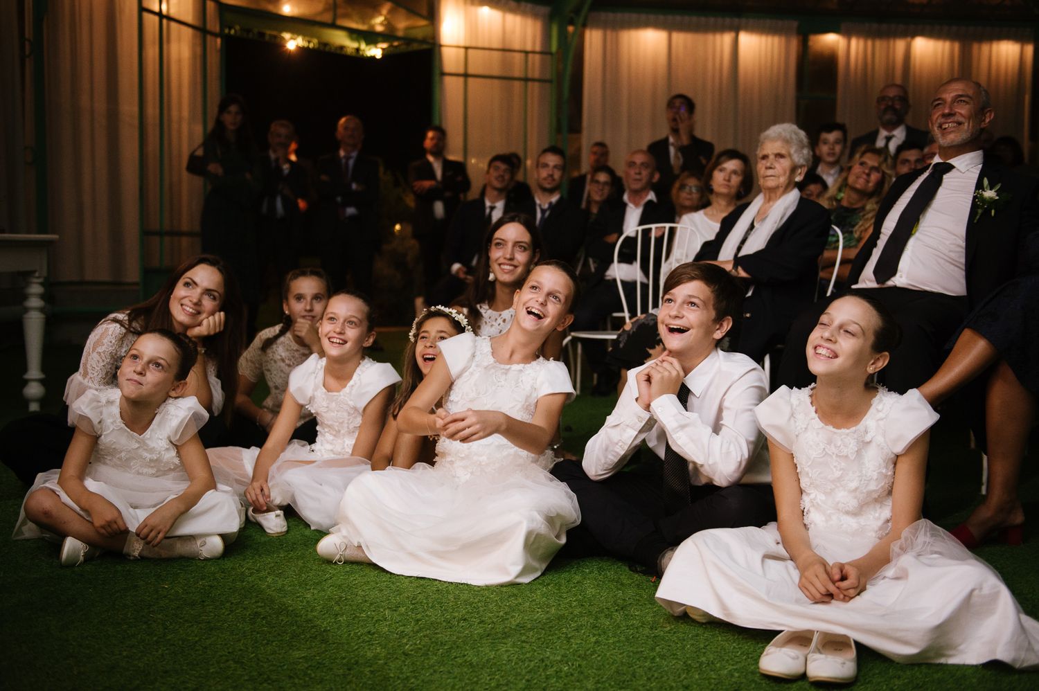 Group of young flower girls in white dresses sitting on grass laughing at a wedding reception.