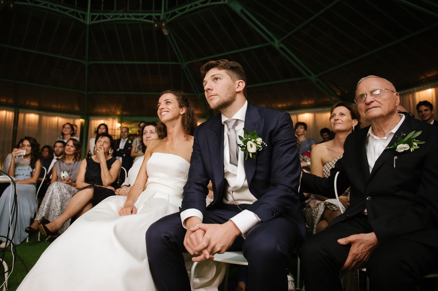 Bride and groom holding hands at wedding ceremony, seated with elderly guest, guests applauding in background.