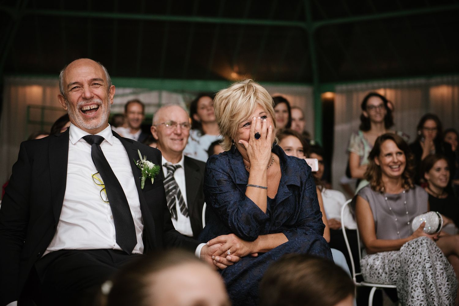 Guests laughing and smiling joyfully at a wedding reception, seated in white chairs in a decorated venue.