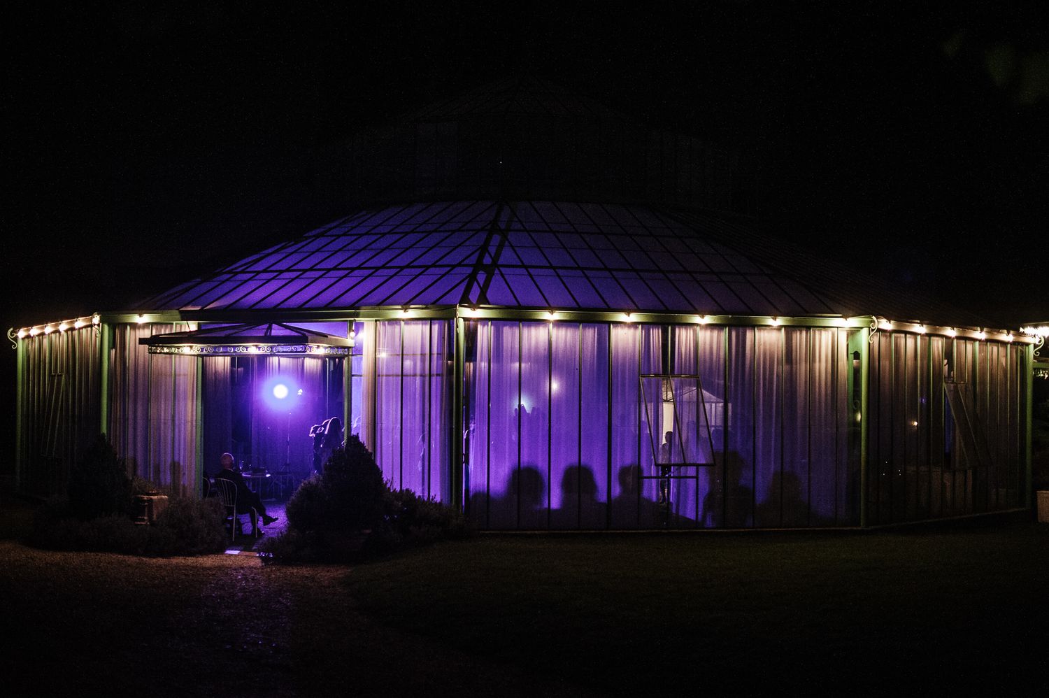A wedding venue glows with purple lights at night, with string lights adorning the exterior of an elegant pavilion.