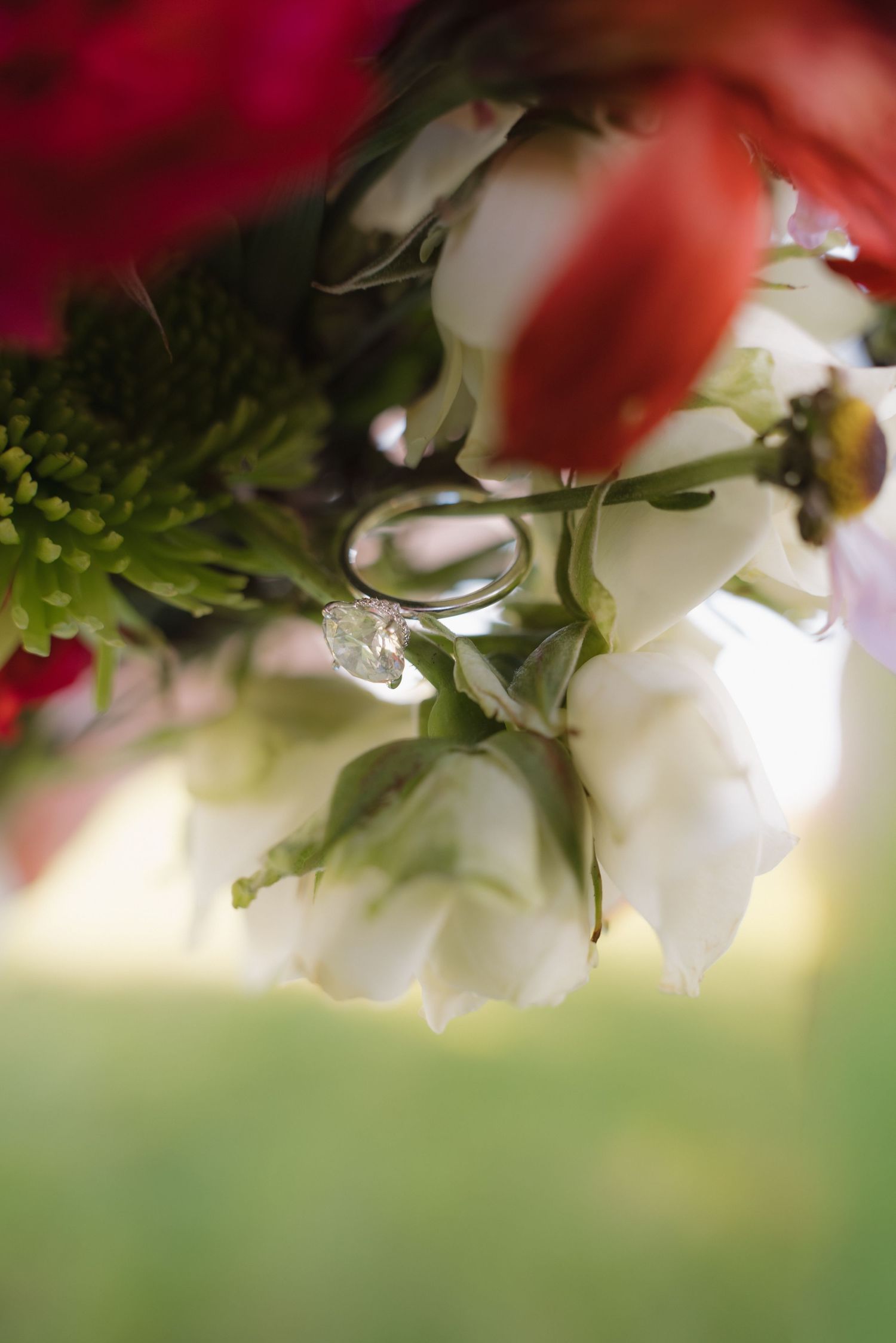 Wedding rings nestled among white, red, and green flowers in a bridal bouquet, with a sparkling diamond engagement ring.
