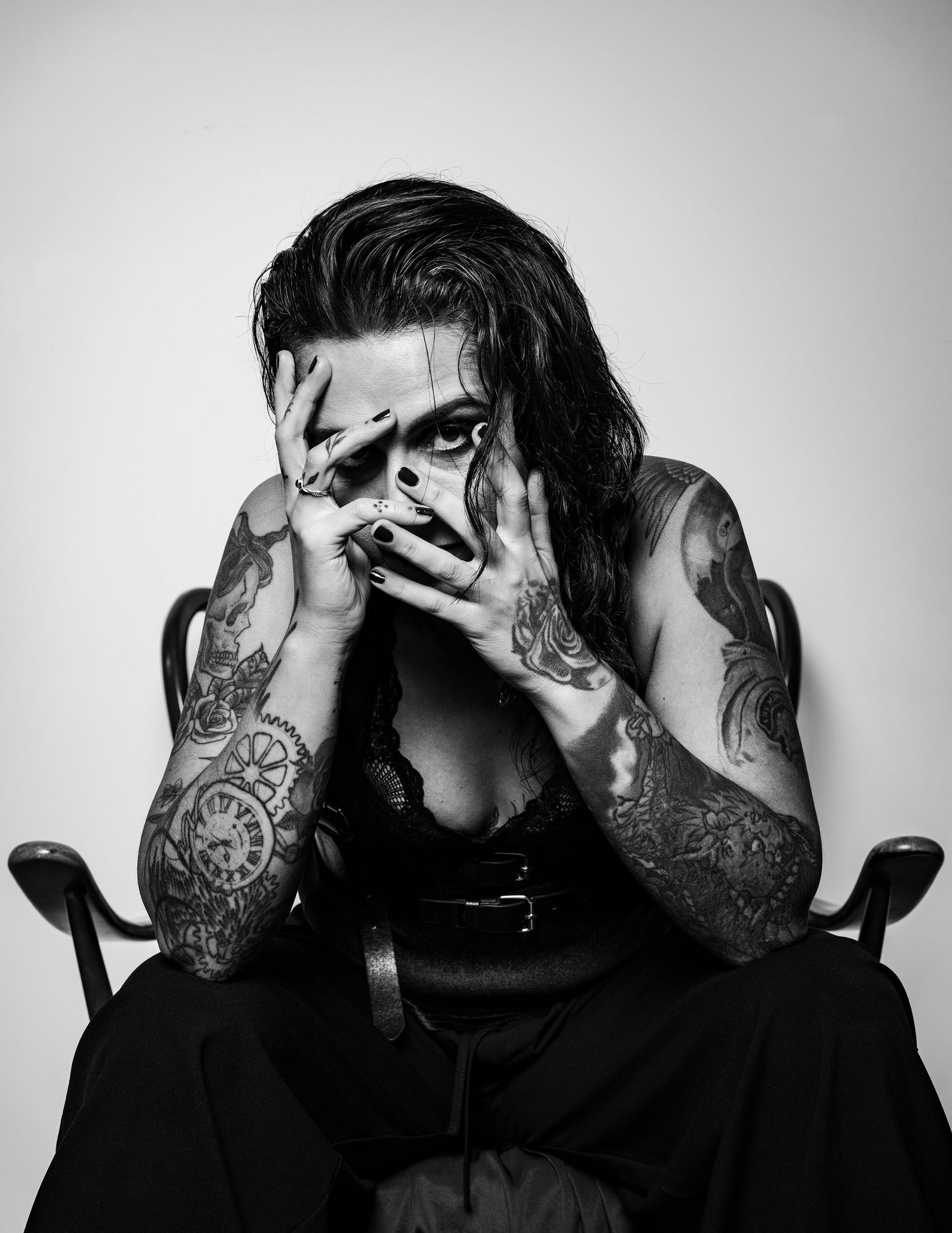 Black and white portrait of a tattooed woman covering her face with her hands, seated in a wooden chair, emotional editorial style