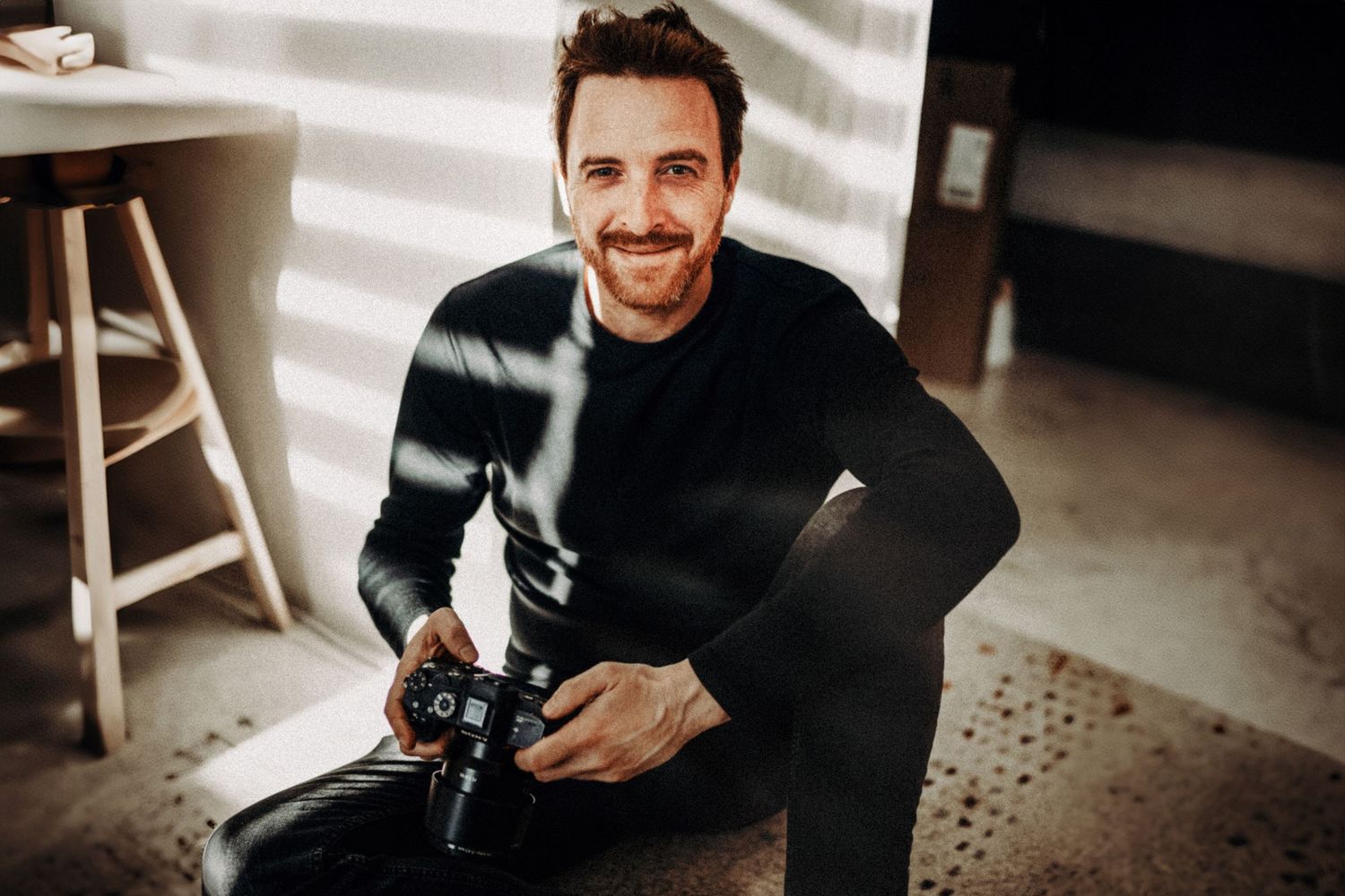 Black and white photo of a smiling young man in a t-shirt leaning forward in a candid, relaxed pose.