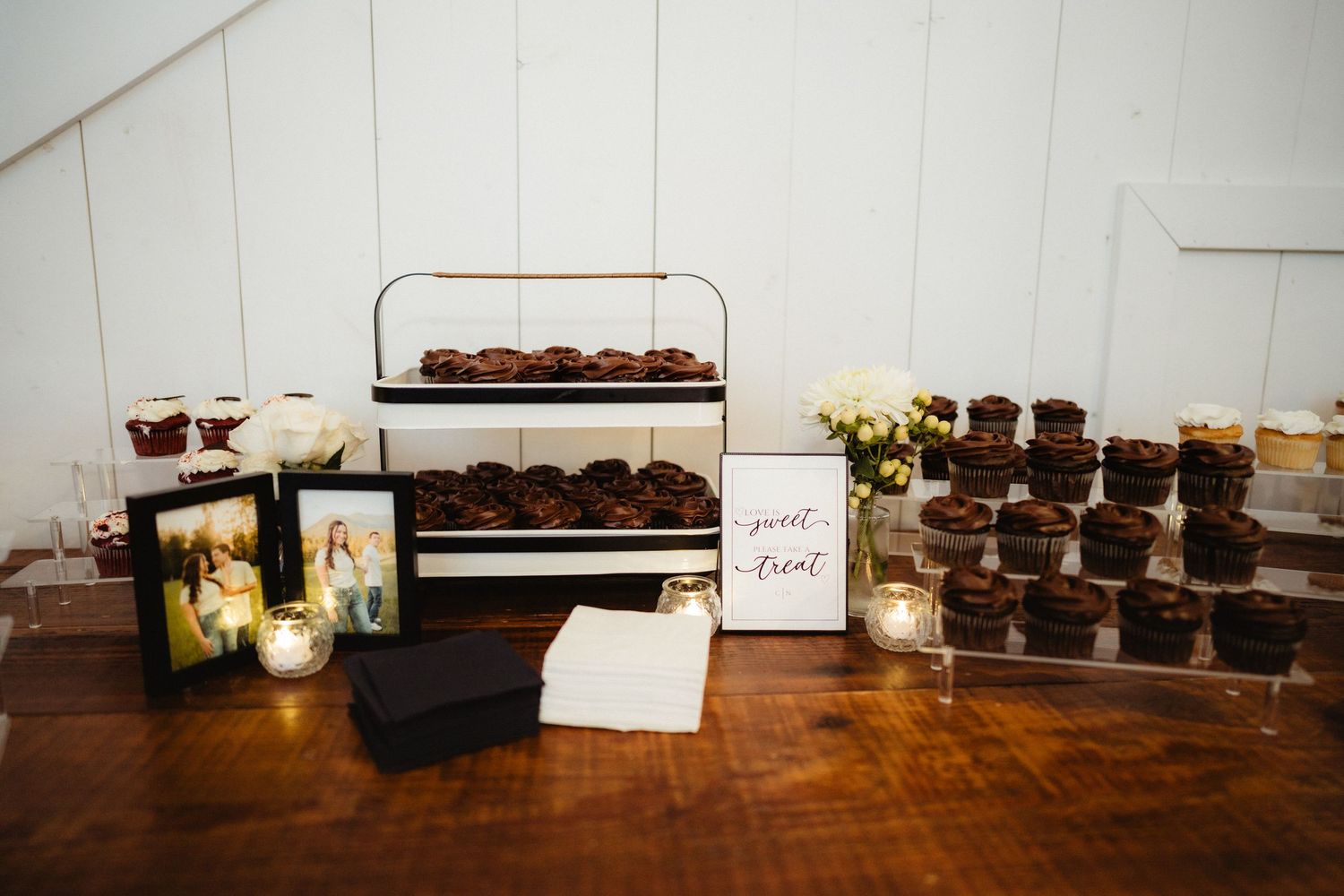 Elegant wedding dessert table with chocolate cupcakes, candles, framed photos, and a sweet treat sign on a rustic wood table.