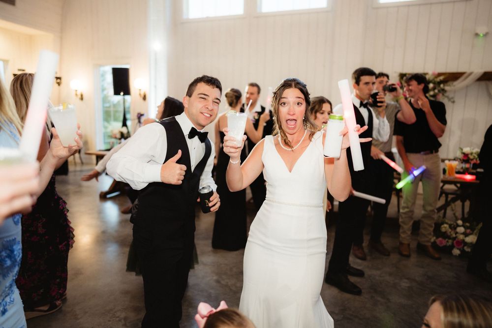 Bride and groom celebrate joyfully with glow sticks at their wedding reception surrounded by cheering guests.