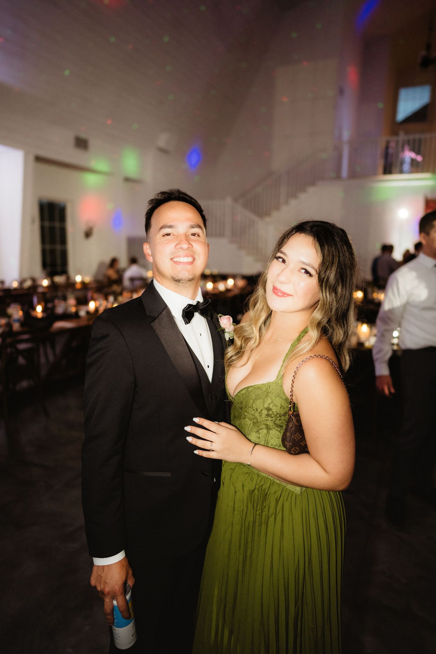 A couple poses at a wedding reception, man in tuxedo and woman in green gown, with colorful lights in background.