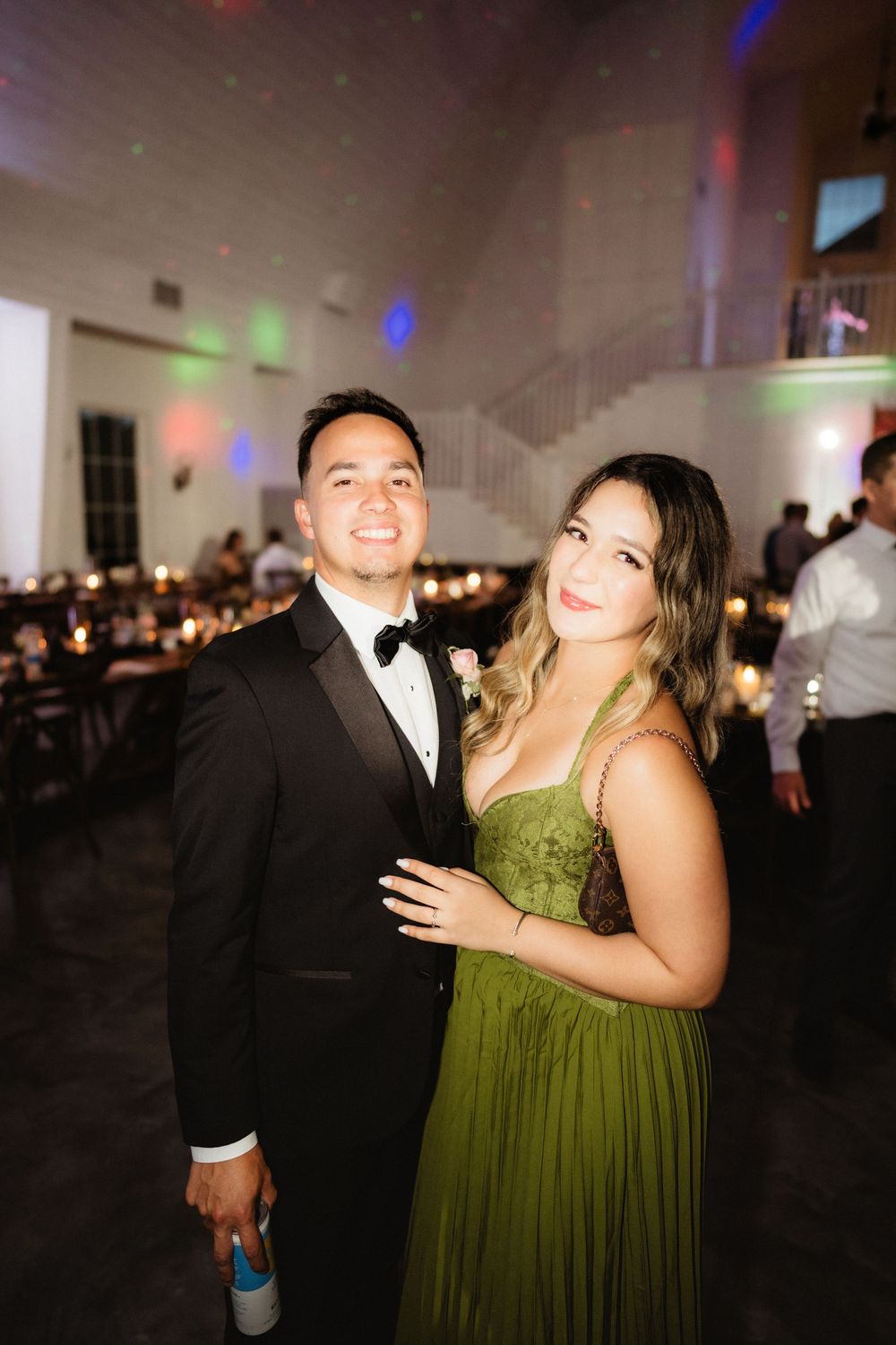 A couple poses at a wedding reception, man in tuxedo and woman in green gown, with colorful lights in background.