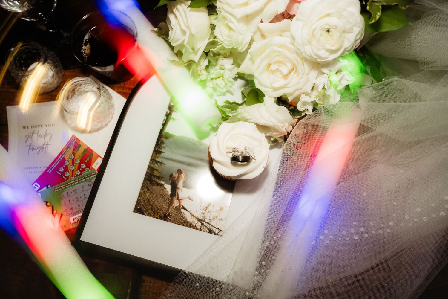 Wedding flat lay with white roses, rings, framed photo, glowing light sticks, and veil on a dark surface.