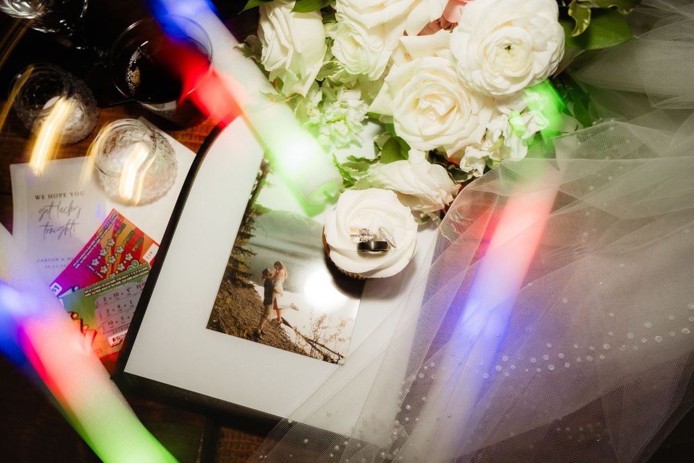 Wedding flat lay with white roses, rings, framed photo, glowing light sticks, and veil on a dark surface.