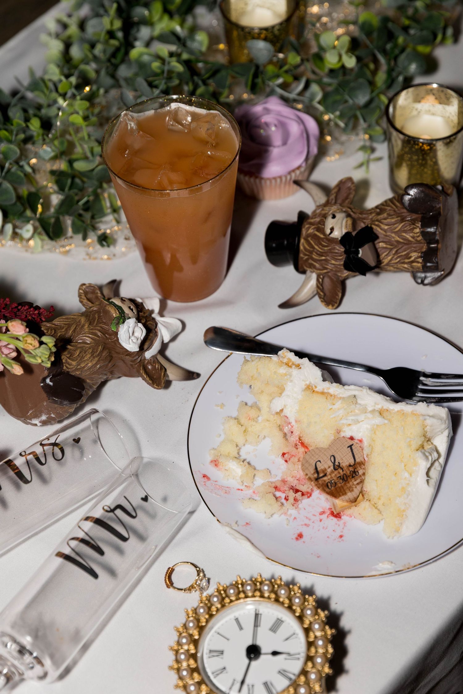 Wedding reception table with cake slice, chocolate figurines, iced tea, cupcake, candles, and vintage clock decor.