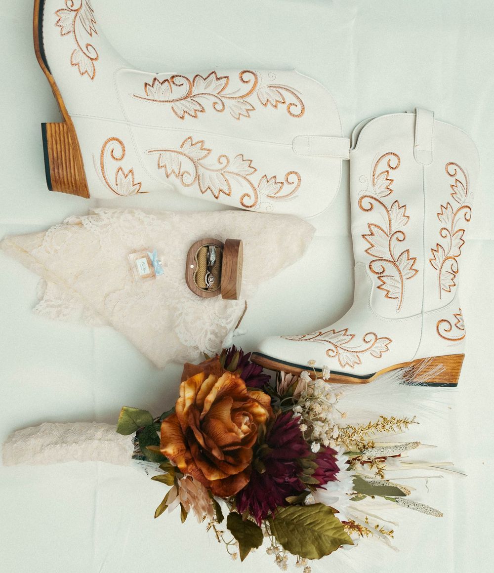 White cowboy boots with gold embroidery displayed with wedding rings and a rustic floral bouquet featuring orange roses and wildflowers.