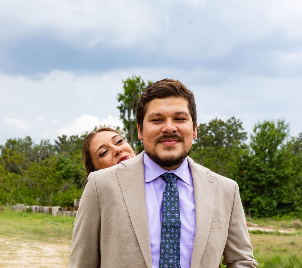 A man in a tan suit and teal tie smiles outdoors while a woman peeks playfully from behind him.