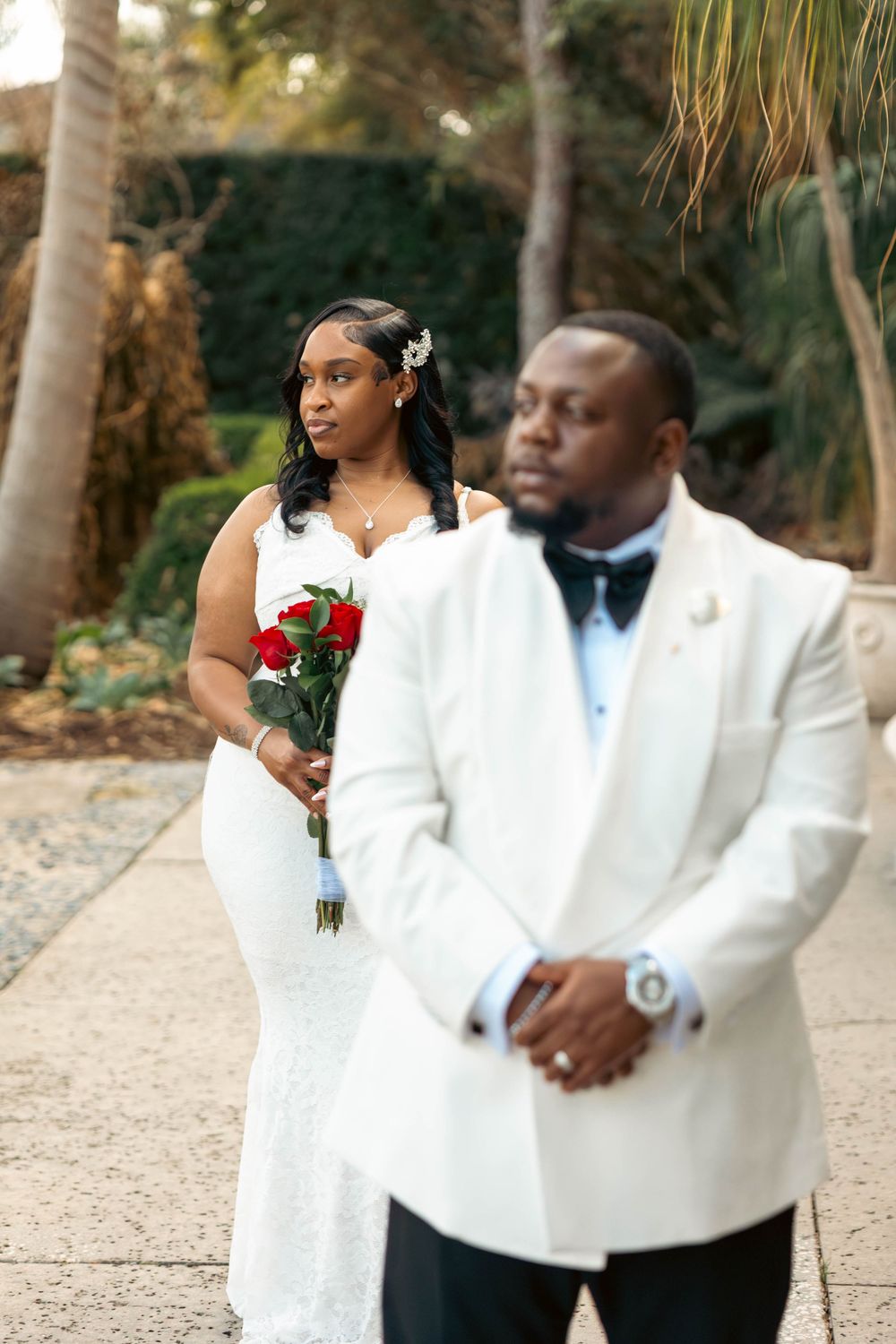 A bride in white lace gown holding red roses stands behind groom in white tuxedo during outdoor wedding photo.